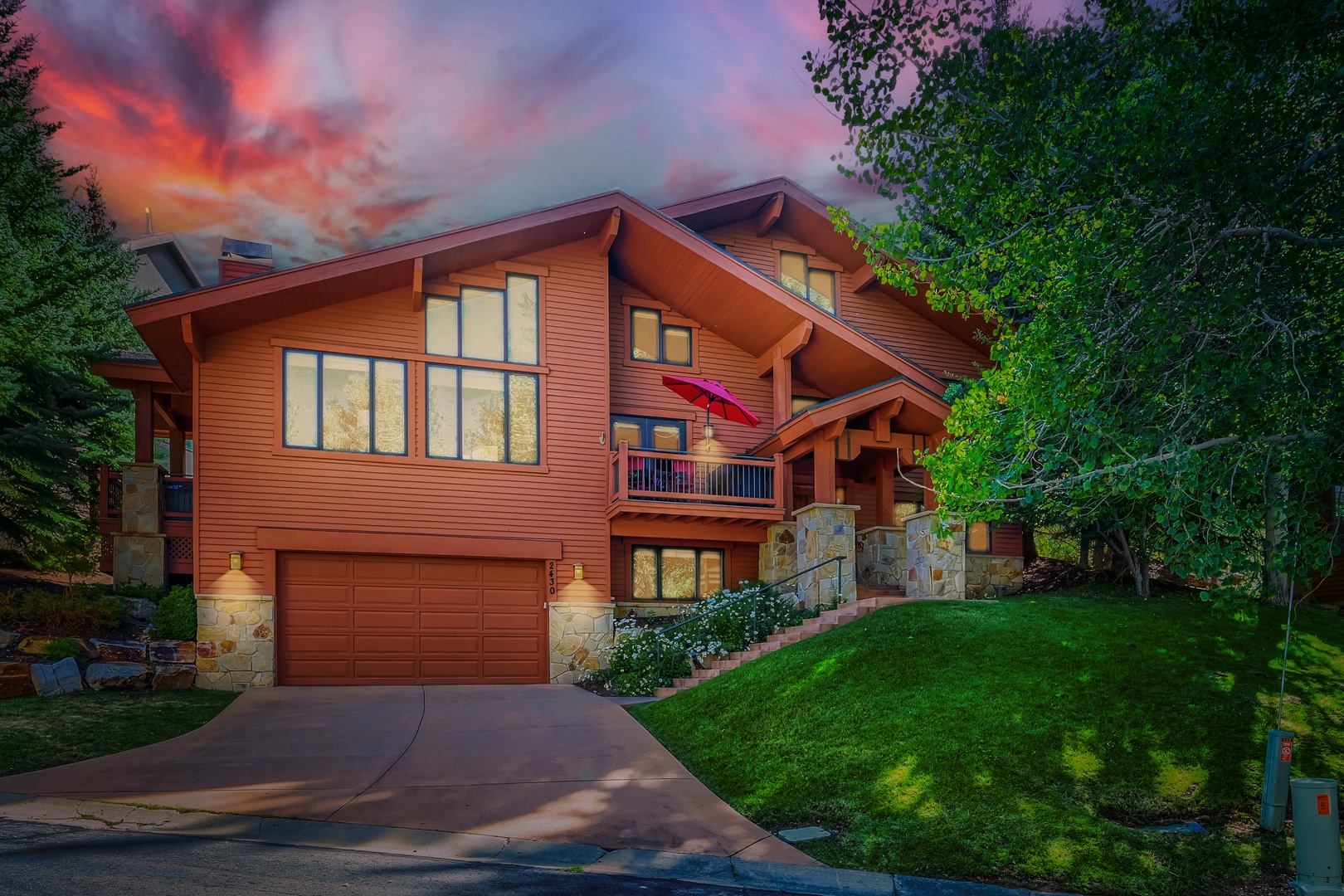 This mountain retreat features warm wood architecture nestled among towering pines, with a welcoming entrance and driveway beneath a spectacular sunset sky.