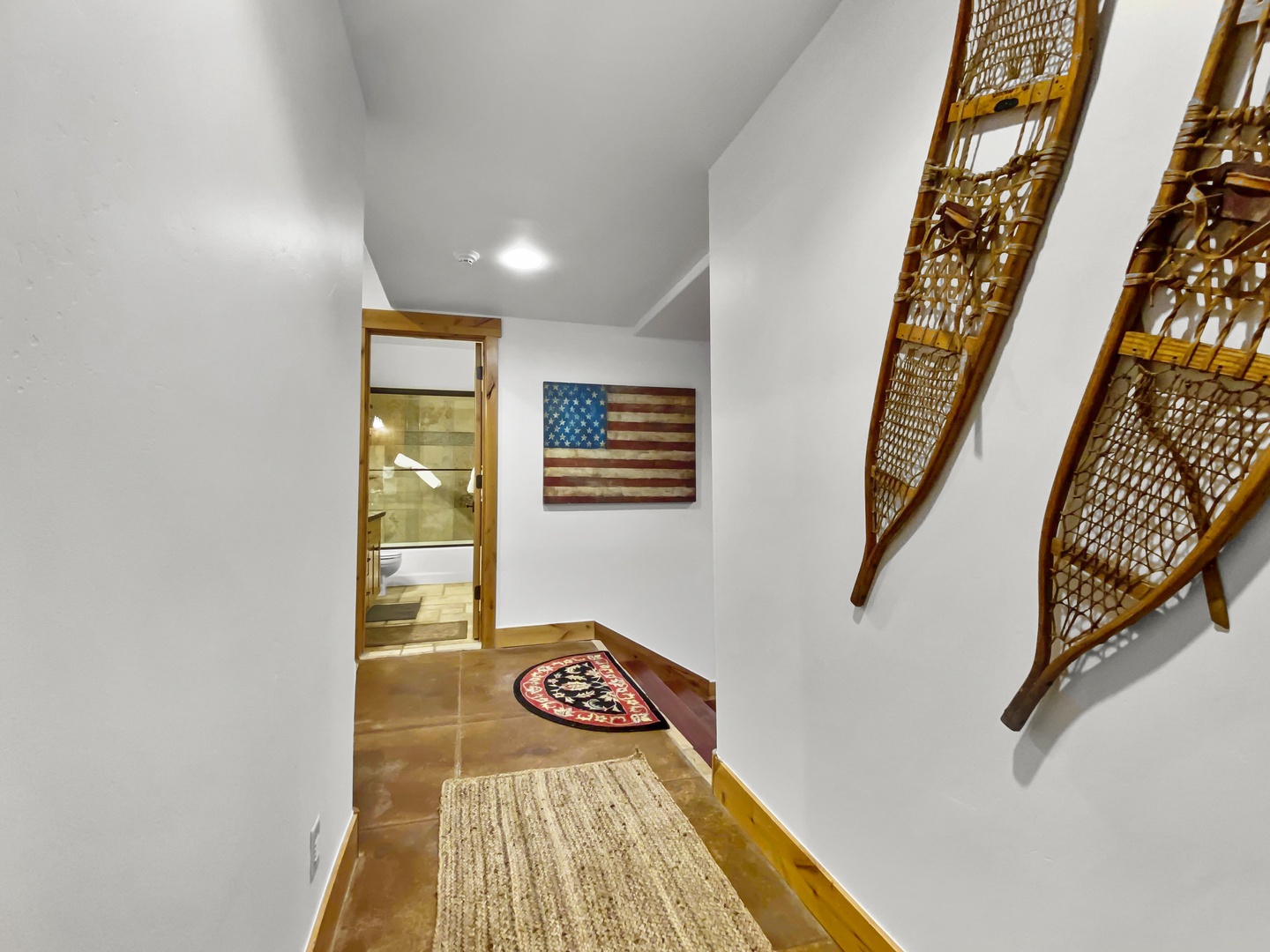 A hallway with a rug on the wooden floor, two snowshoes mounted on the right wall, and an American flag hung at the far end near the entrance to a bathroom. Unit B