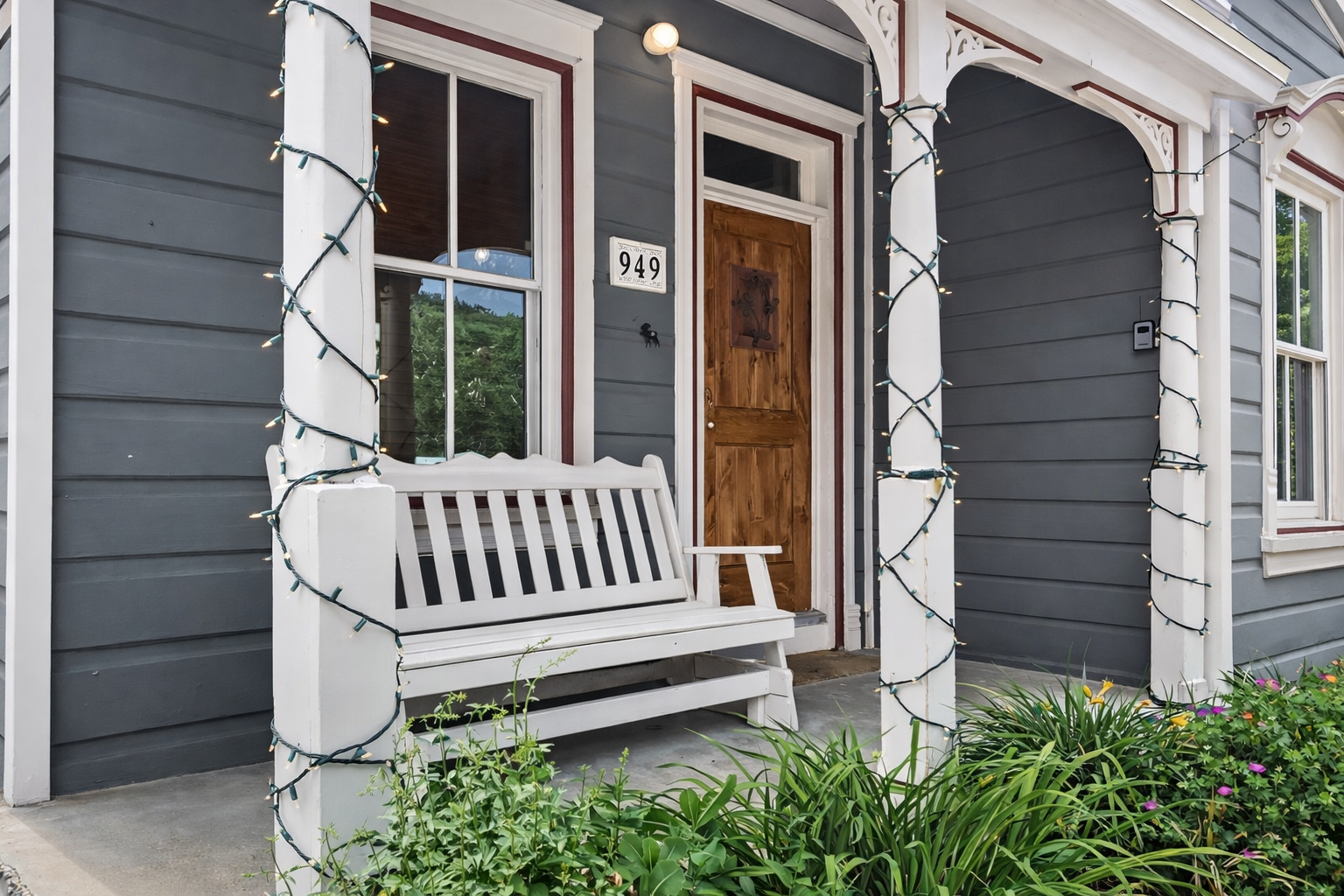 A charming entrance welcomes guests with warm wood doors, classic porch seating, and twinkling lights wrapped around white columns.