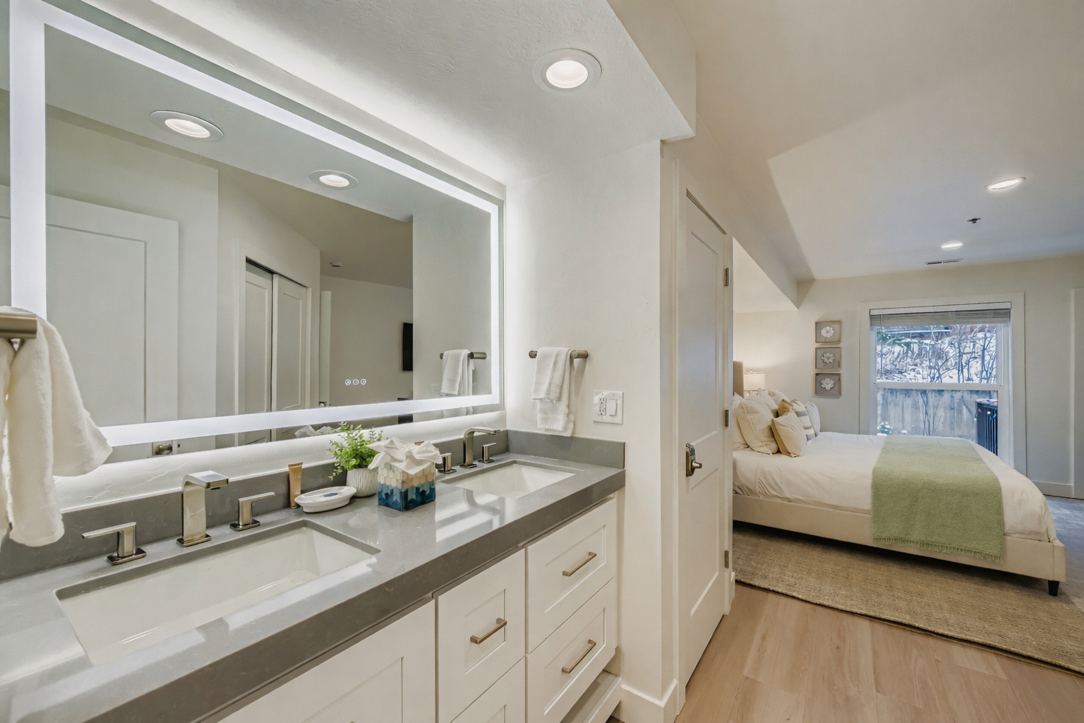 Elegant primary suite with a spa-style double vanity, backlit mirror, and modern finishes. The serene bedroom beyond features a plush king bed and calming tones, offering a bright, comfortable retreat for your Park City stay.