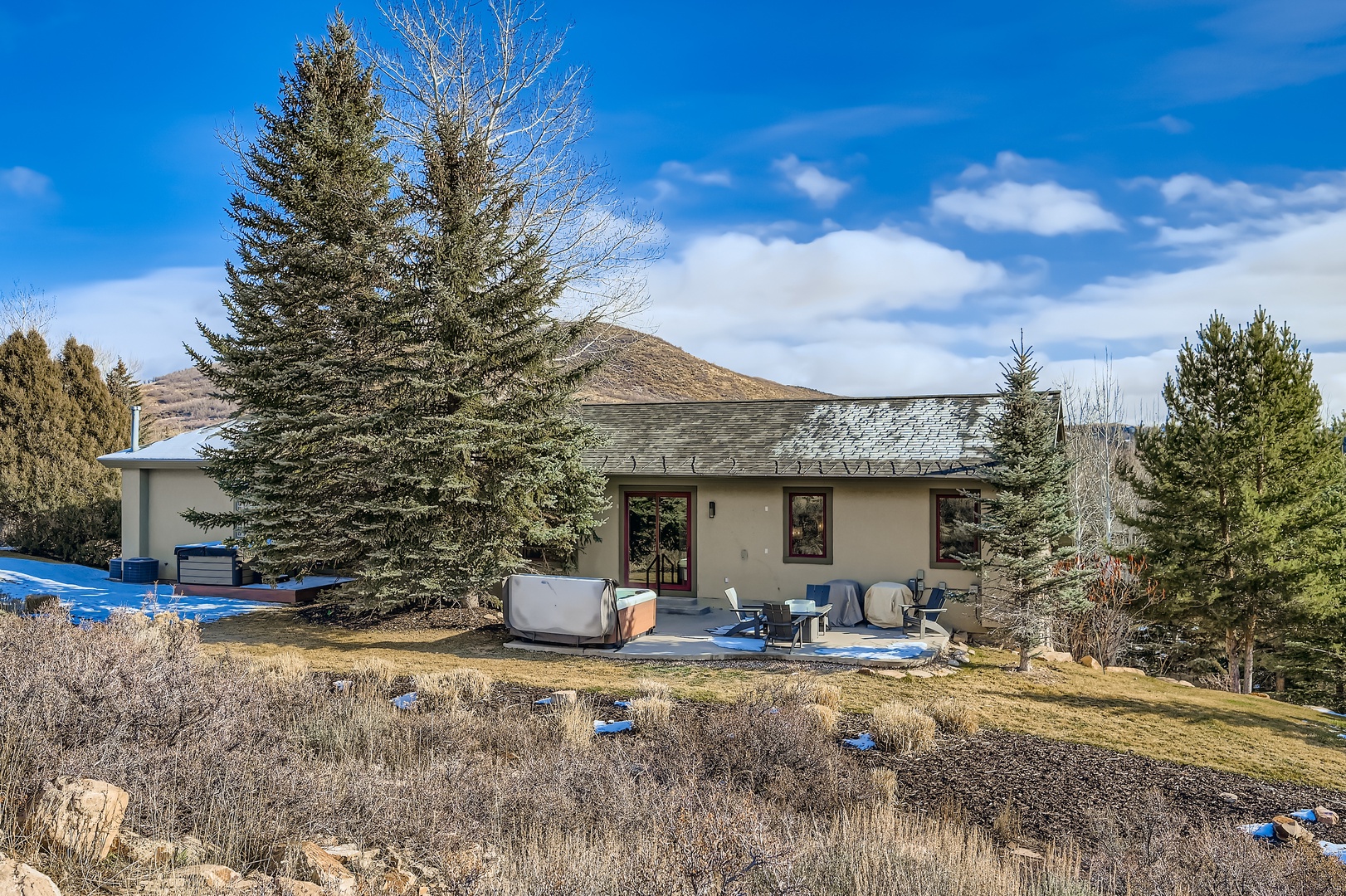 Mountain retreat featuring mature evergreen landscaping, spacious front patio with seating, and rolling hillside terrain under expansive blue skies.