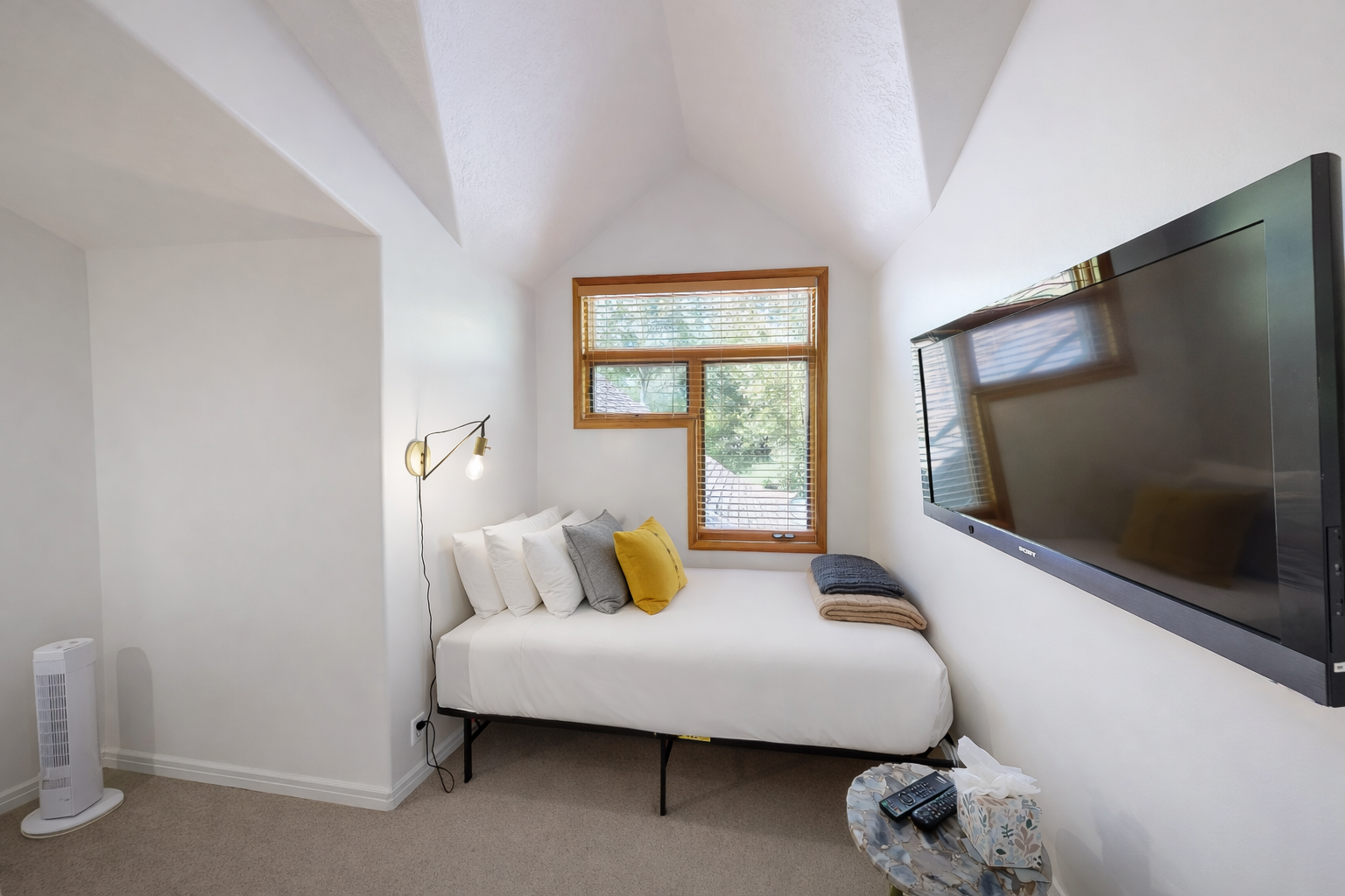 Discover tranquility in this bright, minimalist bedroom featuring a comfortable natural wood bed and a cozy daybed. Large windows offer serene views of the outdoors.