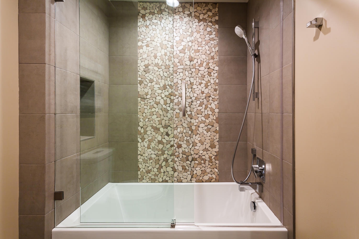 Refresh in this modern bathroom featuring a sleek bathtub, clear glass enclosure, and unique pebble tile accent wall. A stylish space to unwind after a day exploring Park City.