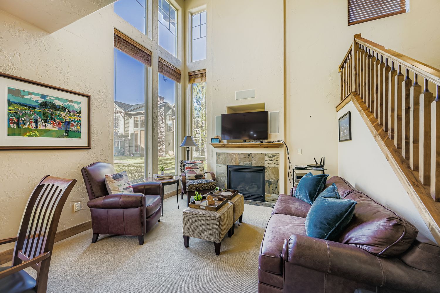 Relax in your spacious living room featuring comfortable leather seating, a cozy fireplace, and soaring ceilings with abundant natural light from tall windows.