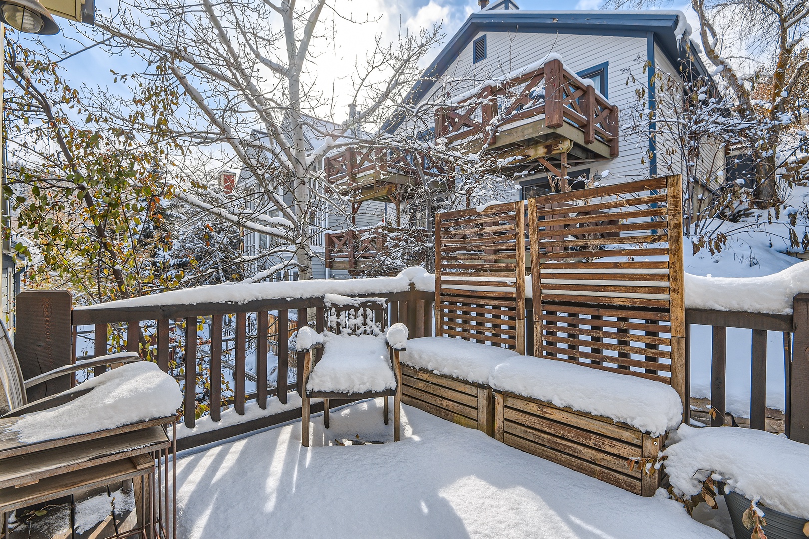 Snow-covered deck with wooden privacy screens leading to a charming two-story structure, surrounded by bare winter trees and pristine snowfall.