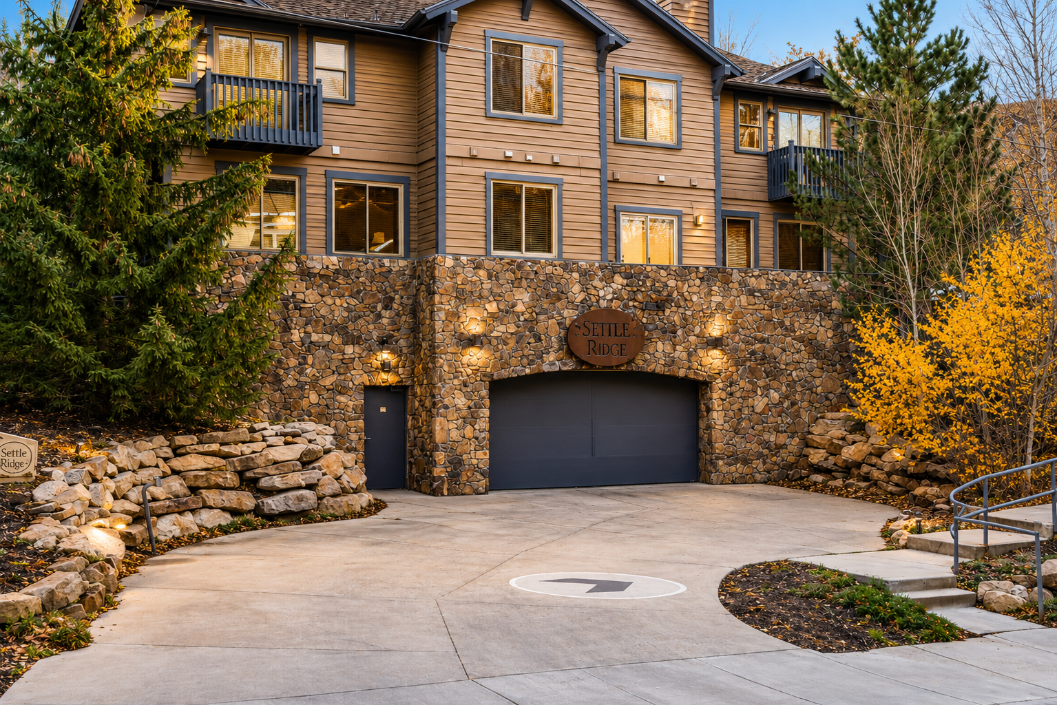 Settle Ridge property exterior features warm brown siding, stone accents, multiple balconies, and a welcoming entrance framed by autumn foliage and landscaped pathways.
