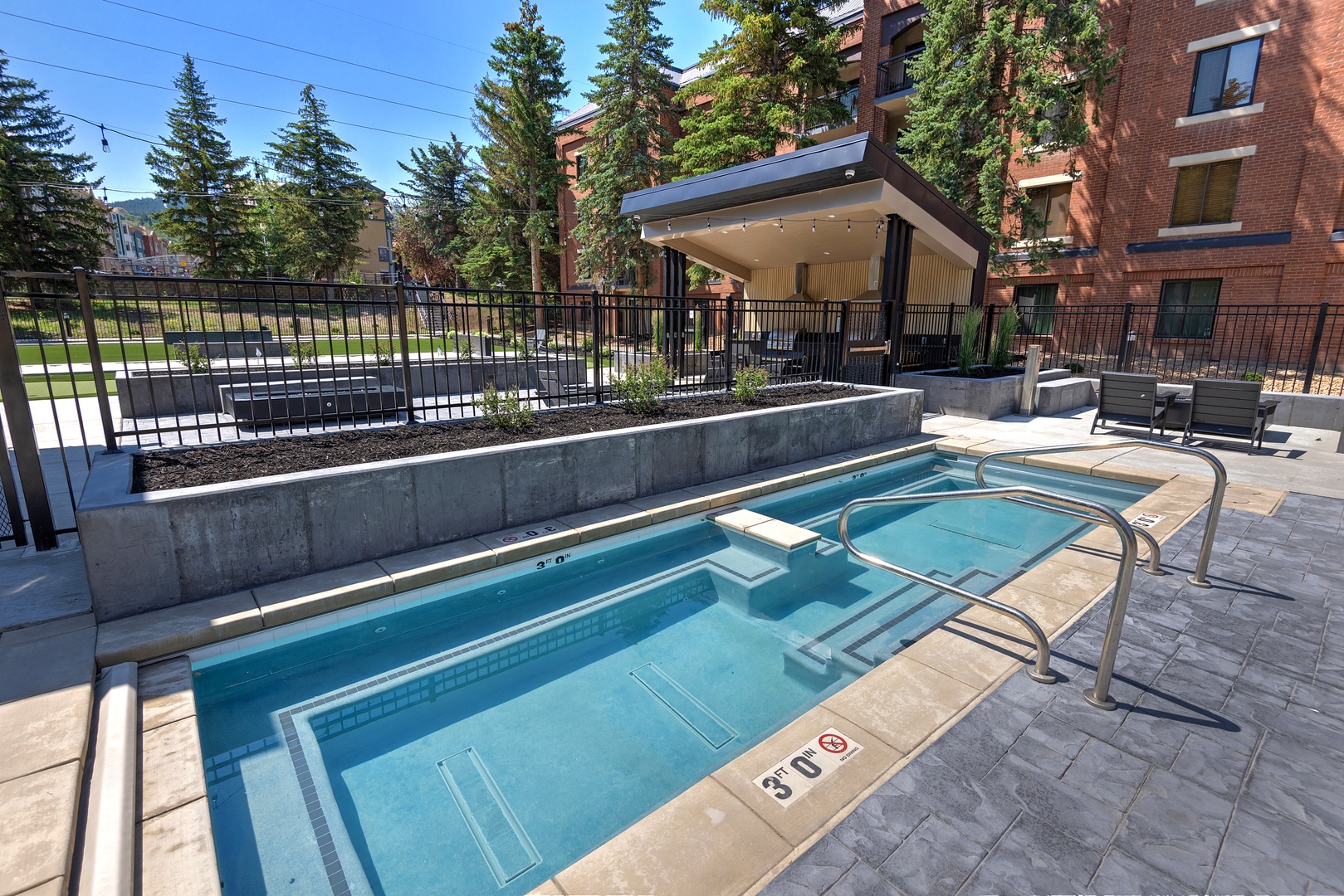 Modern outdoor pool area nestled in a residential setting, with a covered pavilion for relaxation after your swim. Surrounded by mature trees and just steps from the building.