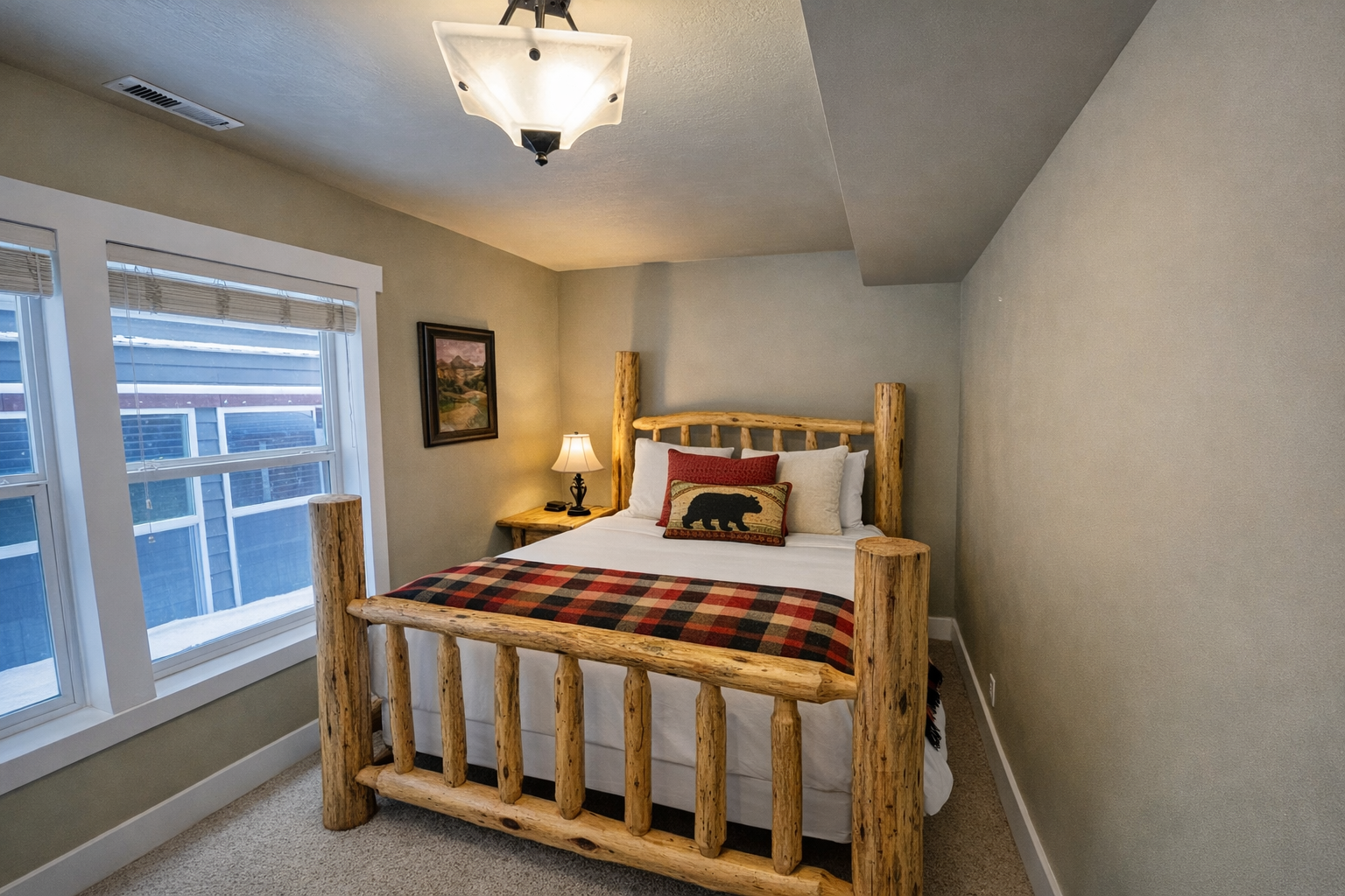 This charming secondary bedroom features a handcrafted log bed frame and cozy buffalo plaid accents. Large windows fill the space with natural light, creating a warm, inviting atmosphere for guests.
