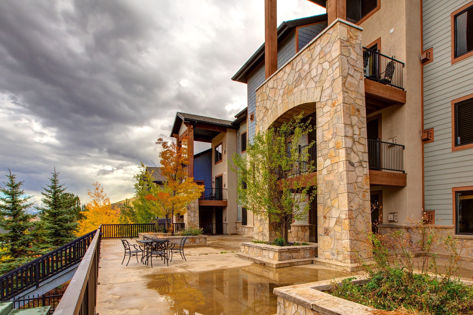 Outdoor patio with a few tables and chairs in front of a multi-story stone and wood building. Trees with autumn foliage surround the area. Overcast sky with grey clouds in the background.