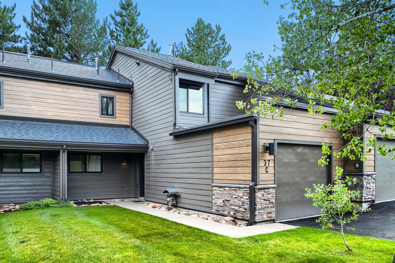 Modern two-story townhome nestled among mature pines, with a fresh lawn, private garage, and contemporary stonework accents welcoming visitors at the entrance.