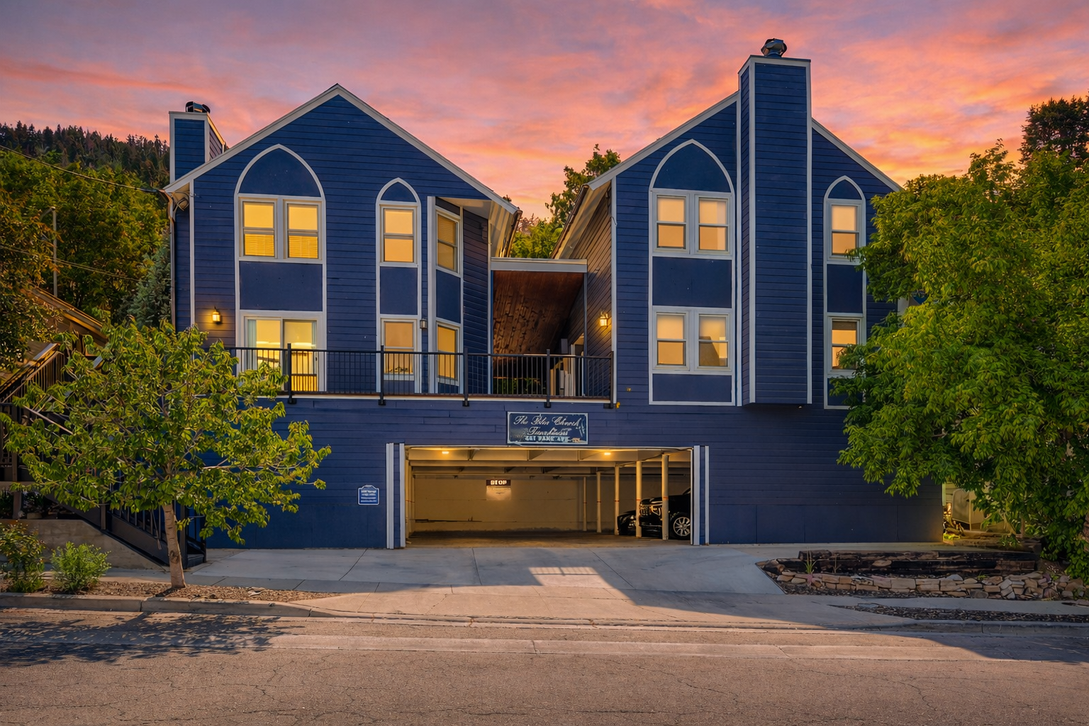 Striking blue lakeside retreat with warm windows glowing at sunset, nestled among lush trees in a peaceful neighborhood setting.