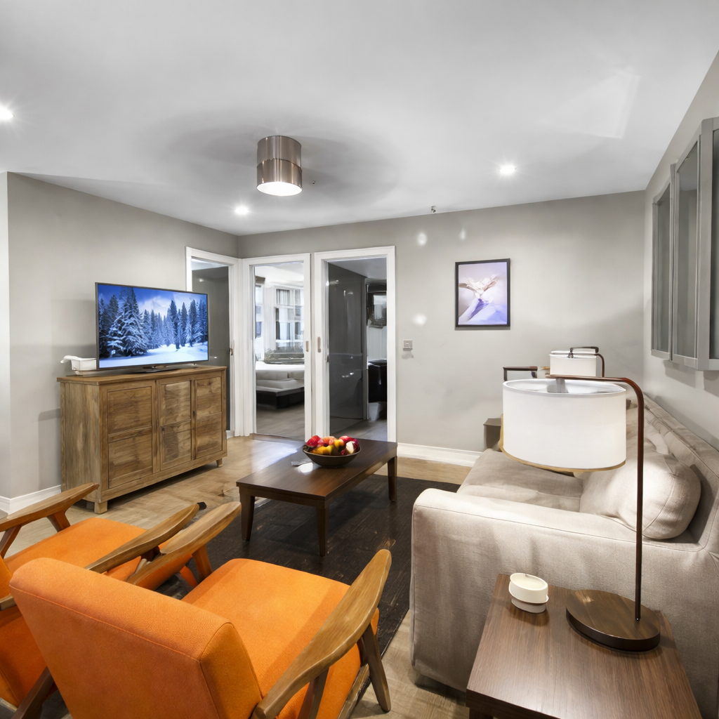 Cozy modern living room featuring vibrant orange chairs, a plush sofa, sleek wood furnishings, and a large TV with a snowy scene. Bright, open layout with warm lighting—perfect for relaxing after skiing or exploring Park City. Ideal for families and group