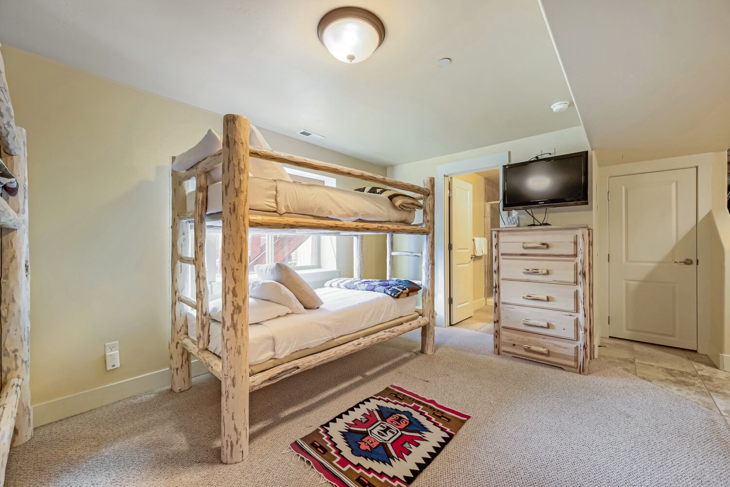 Your cozy bedroom features a charming rustic bunk bed with a natural log frame, offering comfortable sleeping space. Soft carpeting, warm lighting, and a mounted TV create a welcoming mountain retreat perfect for relaxing after your adventures.