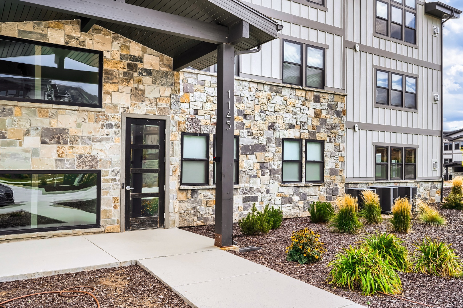 Modern vacation rental with striking stone and metal siding, nestled in a landscaped garden setting with mountain views under bright daytime skies.