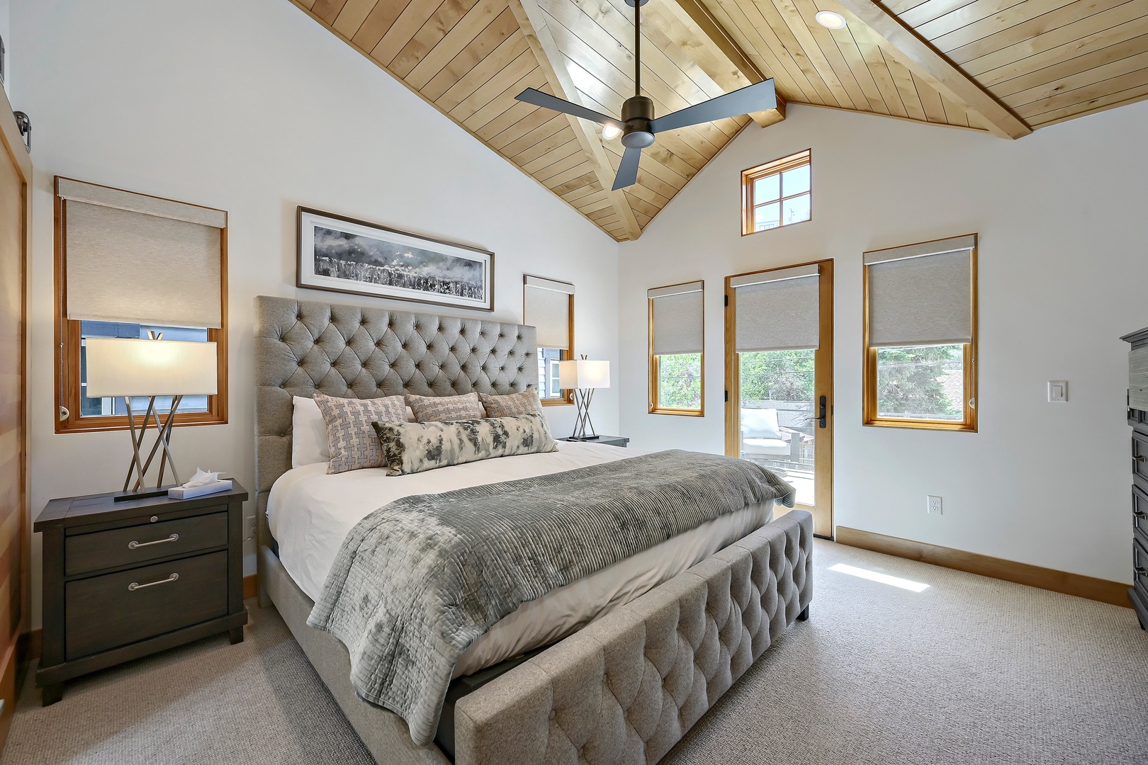 Sink into the plush tufted bed beneath vaulted wood ceilings as natural light streams through multiple windows. Your private retreat features elegant furnishings, warm wood accents, and a balcony door opening to fresh air and views.