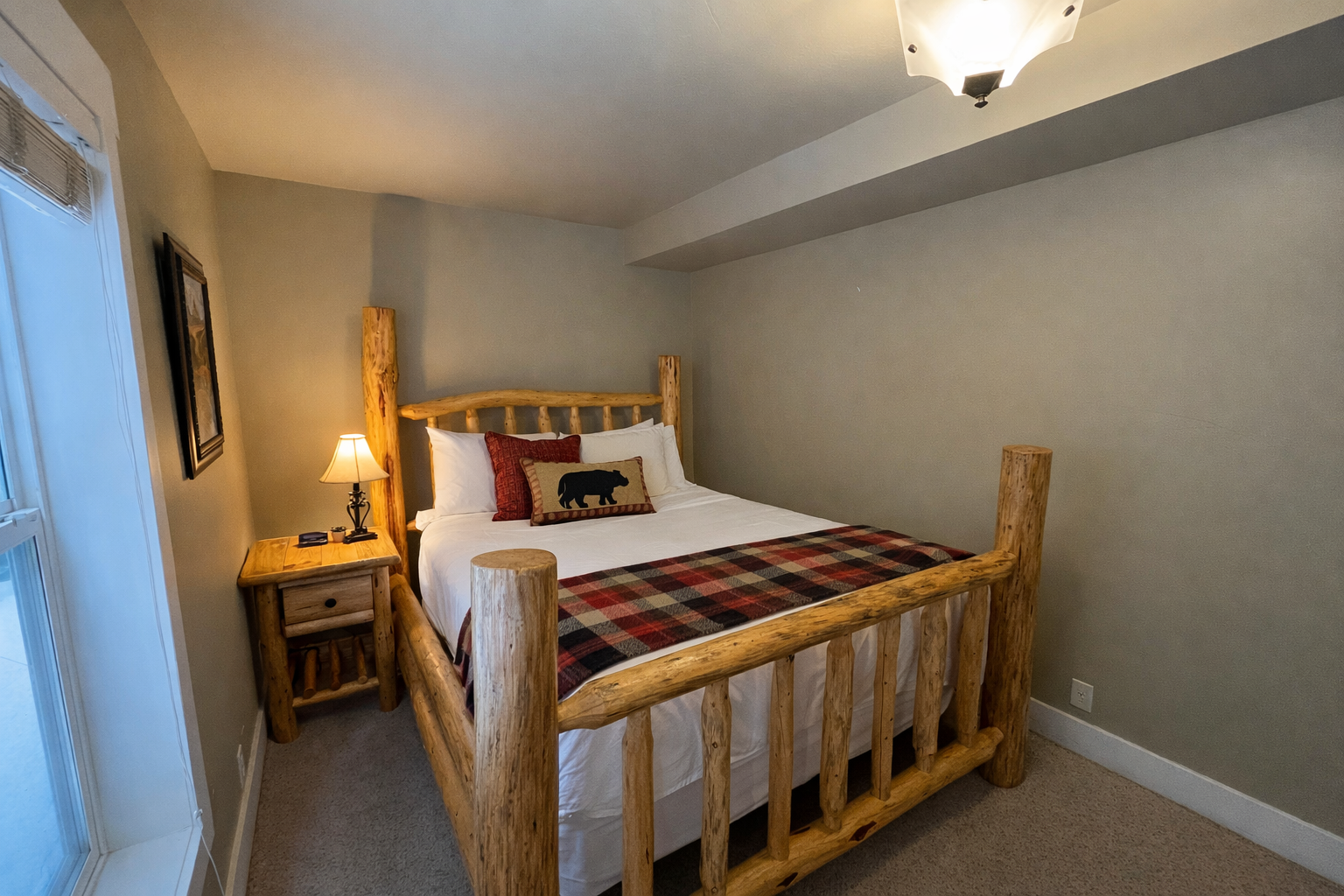 This charming secondary bedroom features a handcrafted log bed frame and cozy buffalo plaid accents. Large windows fill the space with natural light, creating a warm, inviting atmosphere for guests.