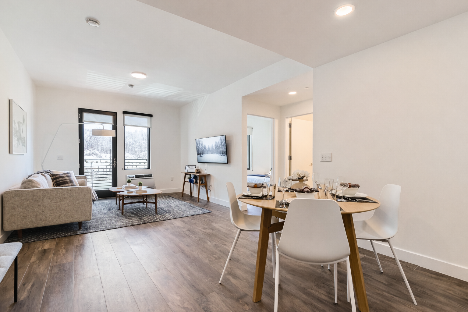 Enjoy this open-concept living area with a dining space for four and a comfortable seating area. The space features modern furniture, wood-look flooring, and large windows with blinds for privacy.