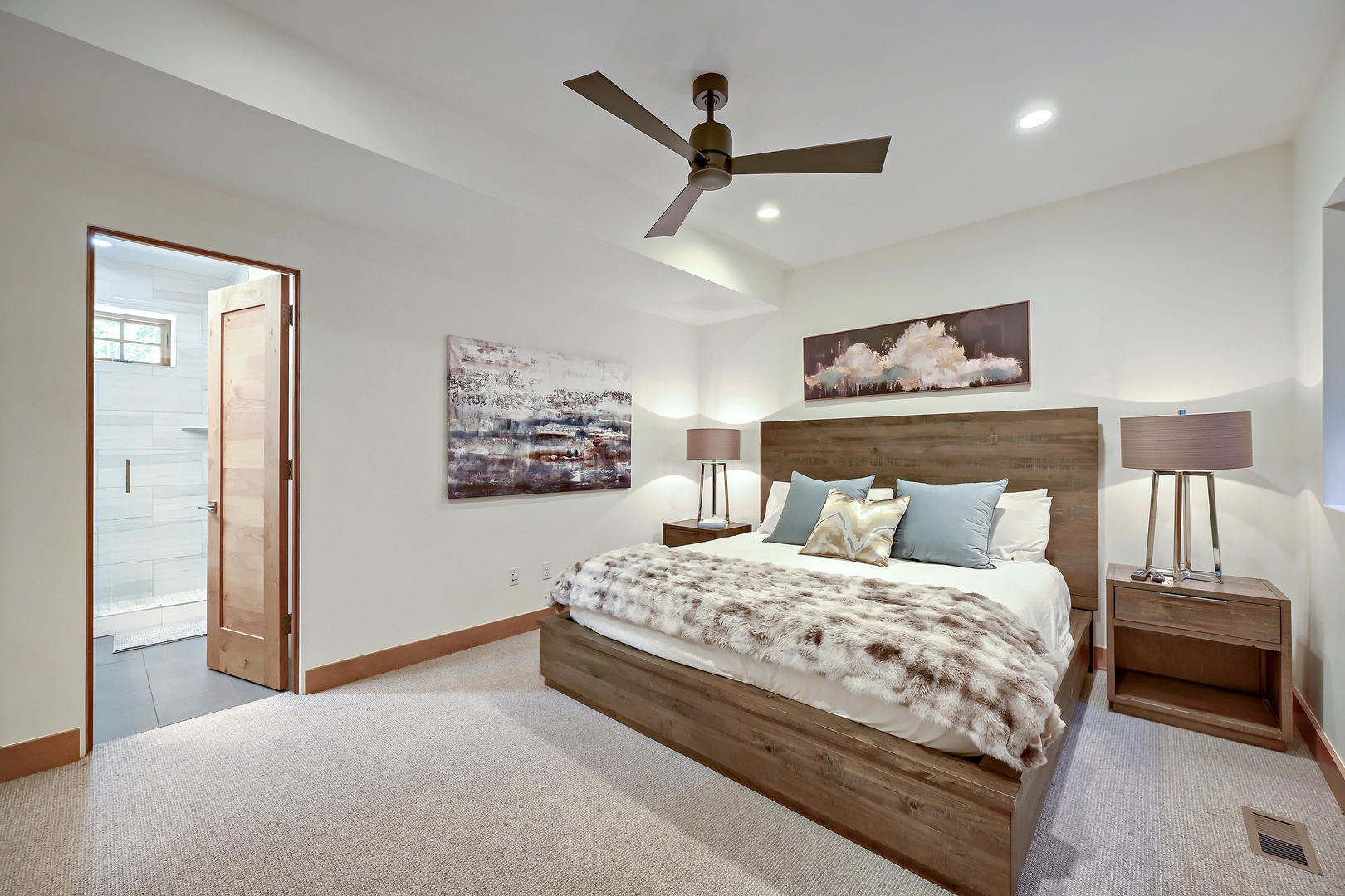 Relax in your serene bedroom featuring a plush bed with soft linens, modern wood furnishings, calming artwork, and a ceiling fan for your comfort.