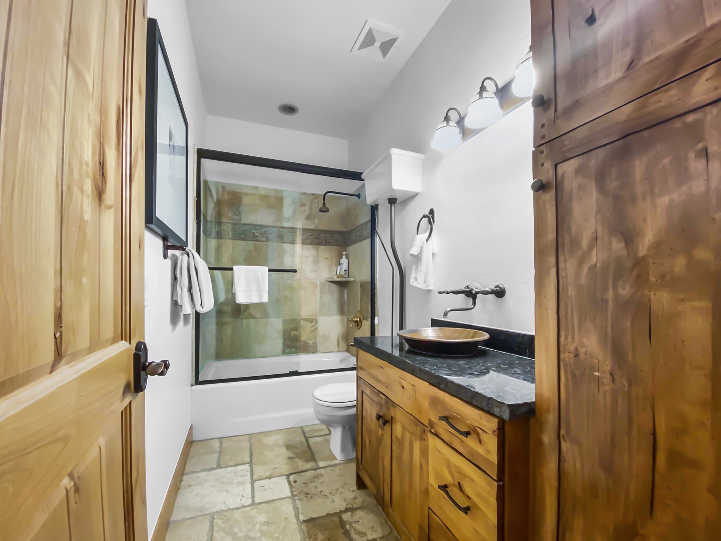 Indulge in this inviting bathroom featuring a rustic wood vanity with a stylish copper sink set in a granite countertop. A tub/shower combo with a sleek glass enclosure completes the serene, natural aesthetic.