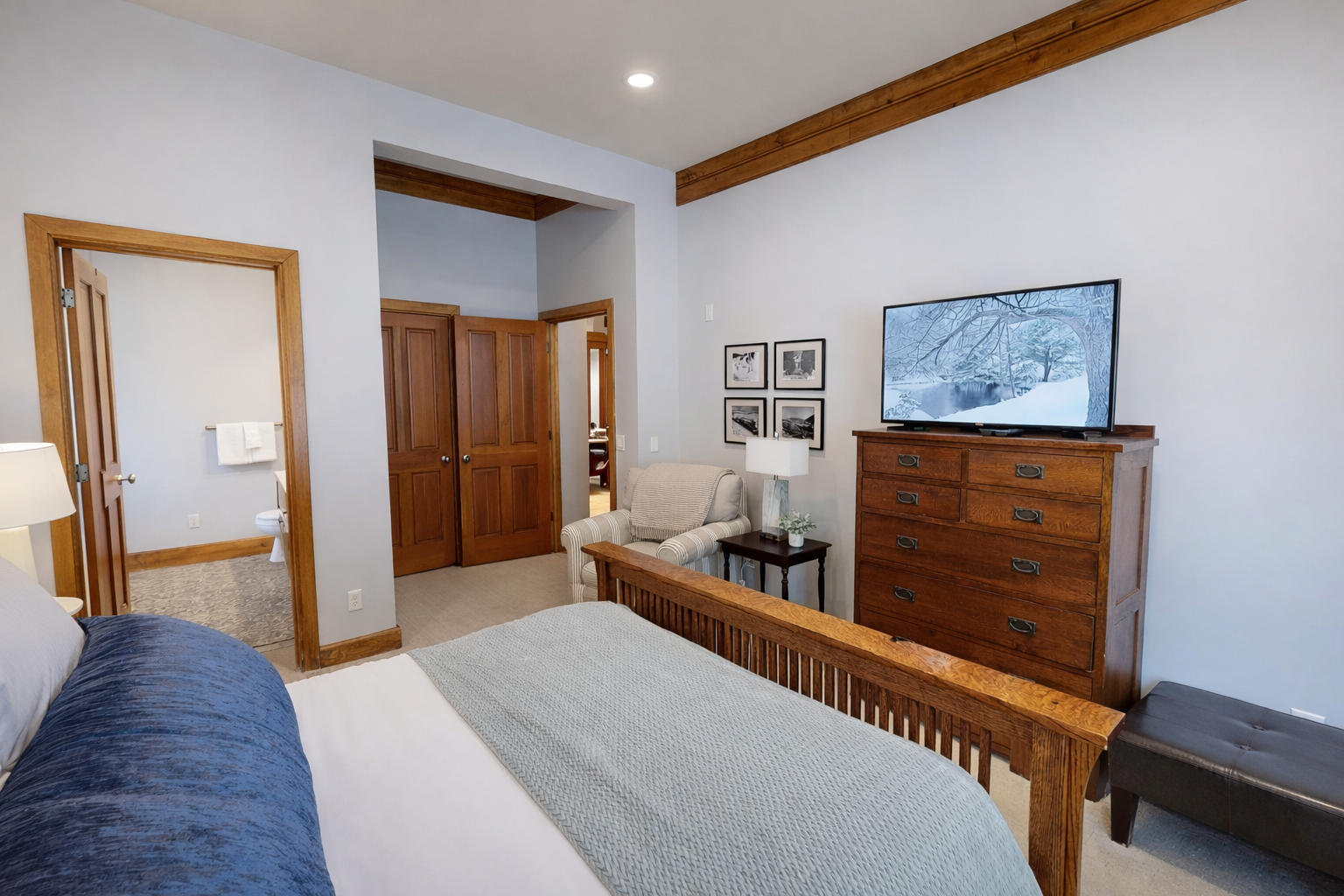 Unwind in the tranquil main floor queen bedroom. Features a plush bed, dark wood furnishings, light wood floors, and large windows with serene forest views for a peaceful stay.