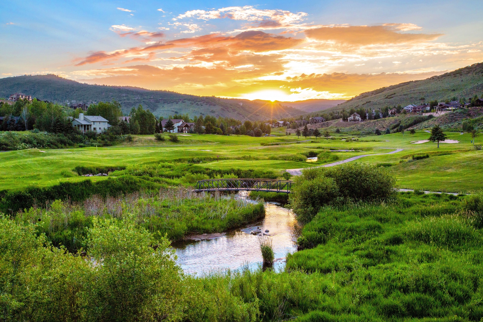 Stunning mountain valley sunset with rolling green hills, meandering stream, and scattered homes creating a peaceful rural landscape.