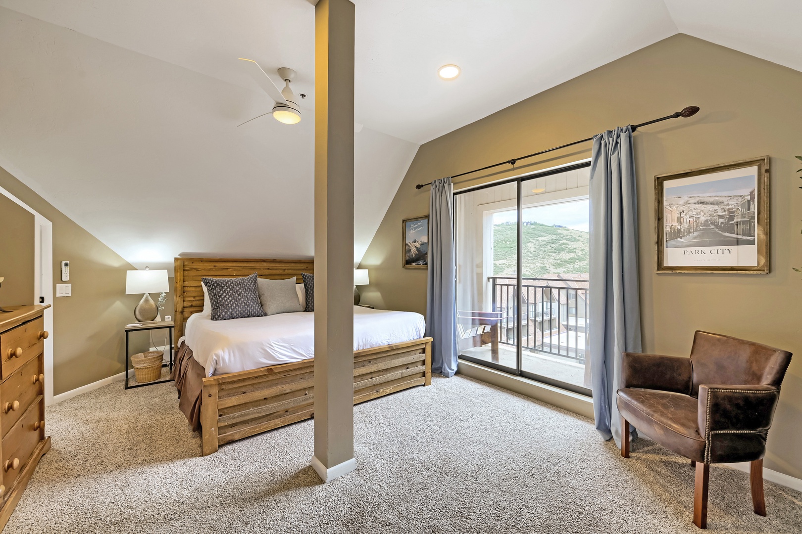 Your inviting bedroom features a cozy bed with warm wood accents, a leather reading chair, and a private balcony framing mountain views—perfect for winding down after your adventures.