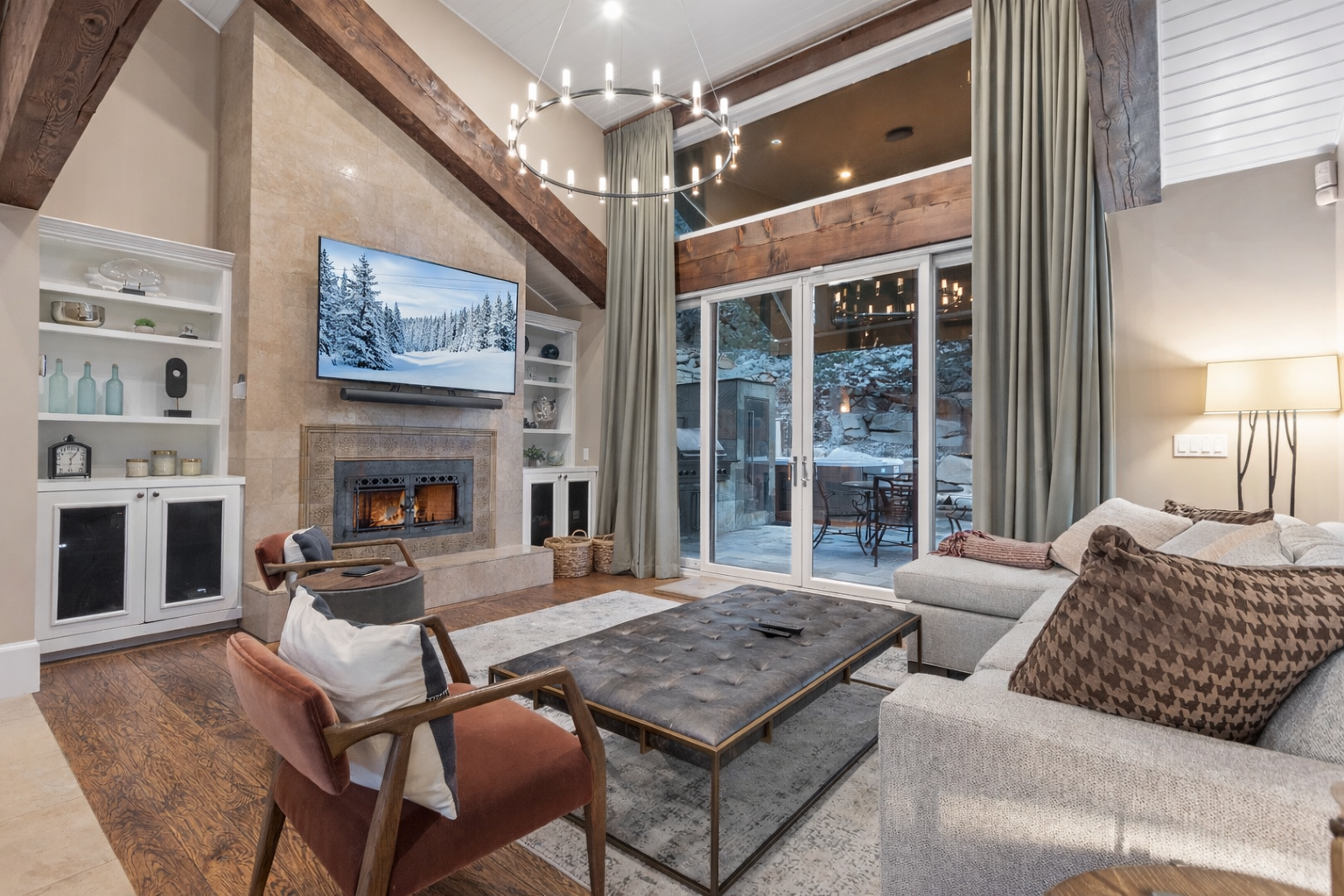 Unwind in this stunning living room with exposed beams, crackling fireplace, and floor-to-ceiling windows revealing snowy mountain views.