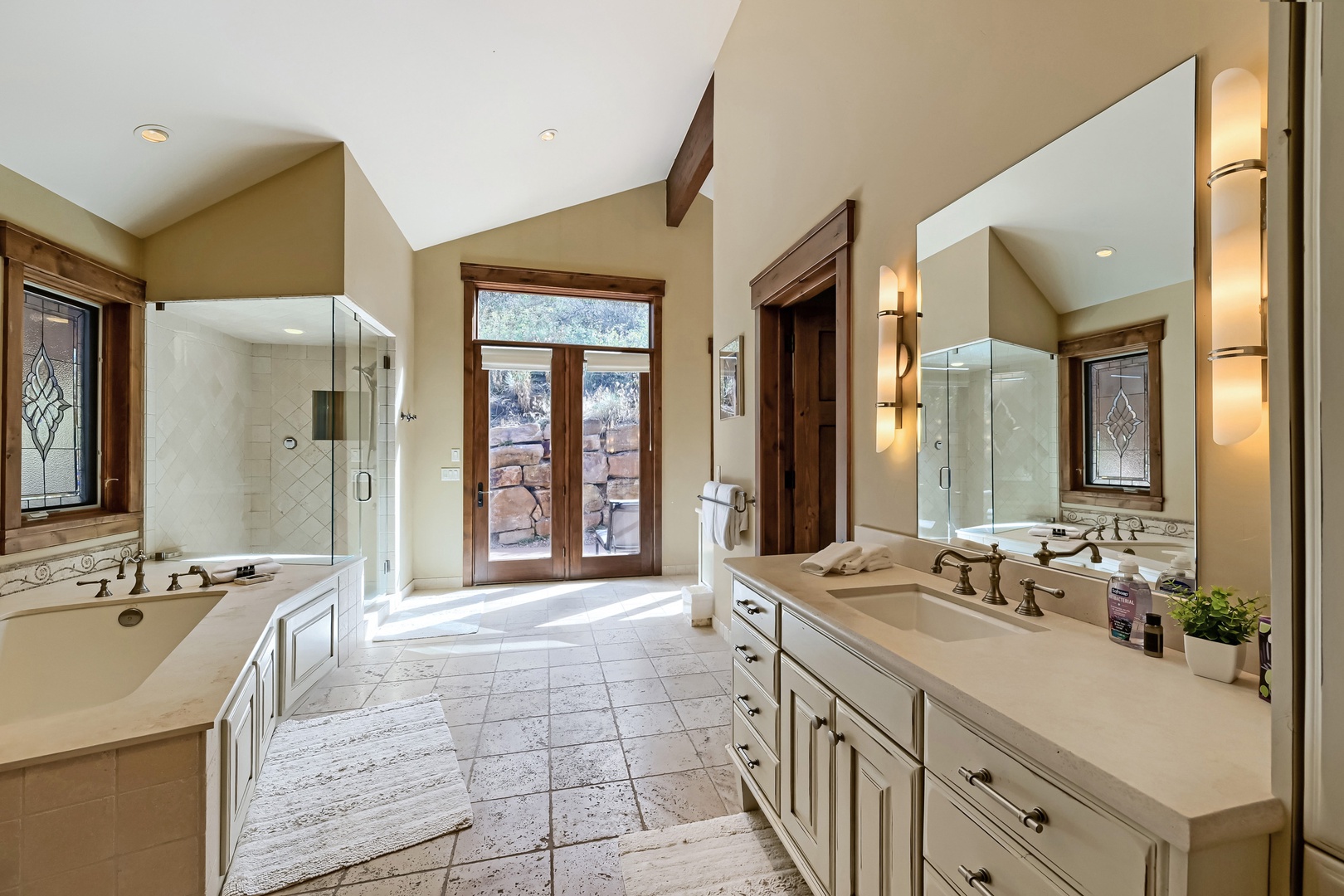 A spacious bathroom designed for relaxation, featuring a deep soaking tub, separate glass-enclosed shower, dual vanities with elegant countertops, and natural light from doors opening to a private outdoor area.