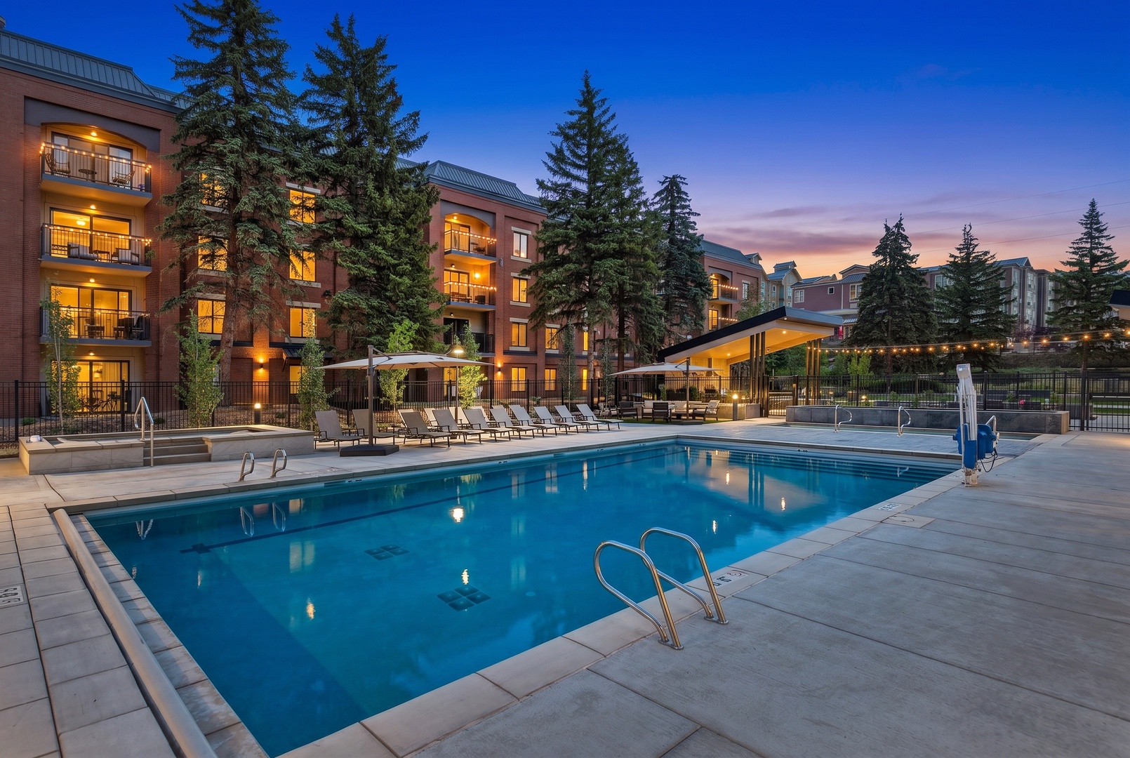 Modern multi-story property with outdoor pool surrounded by towering evergreens, featuring ambient lighting and covered lounge areas as twilight settles over the peaceful mountain setting.