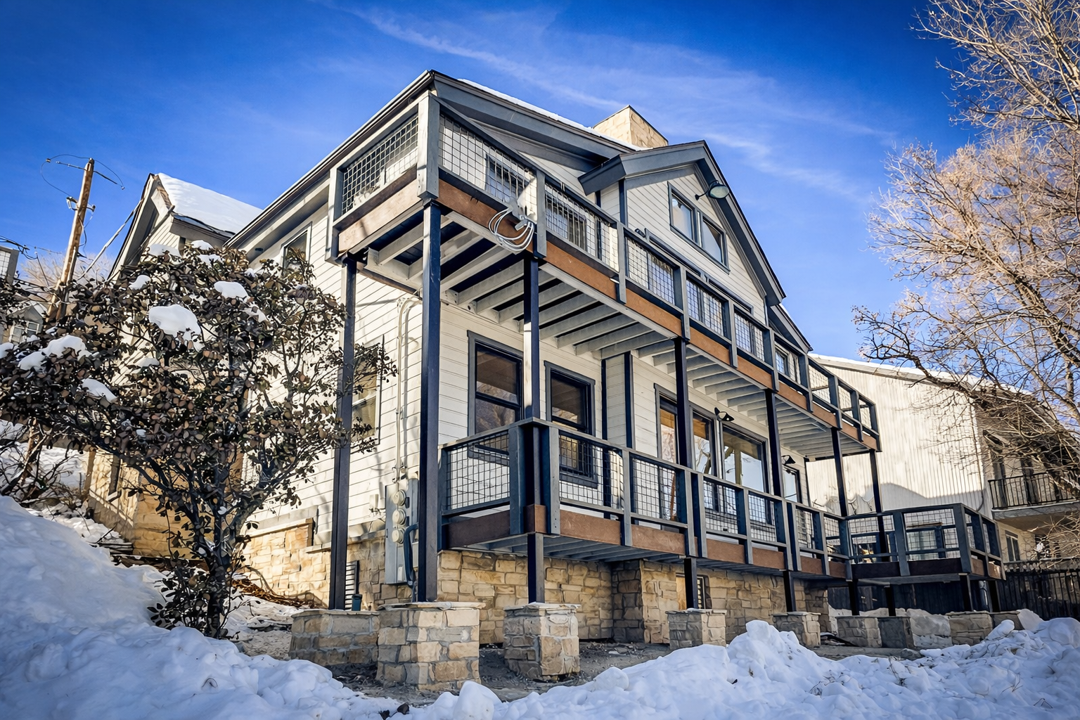 Modern mountain property with stone foundation and covered decks, nestled in a snowy winter setting with bare trees.