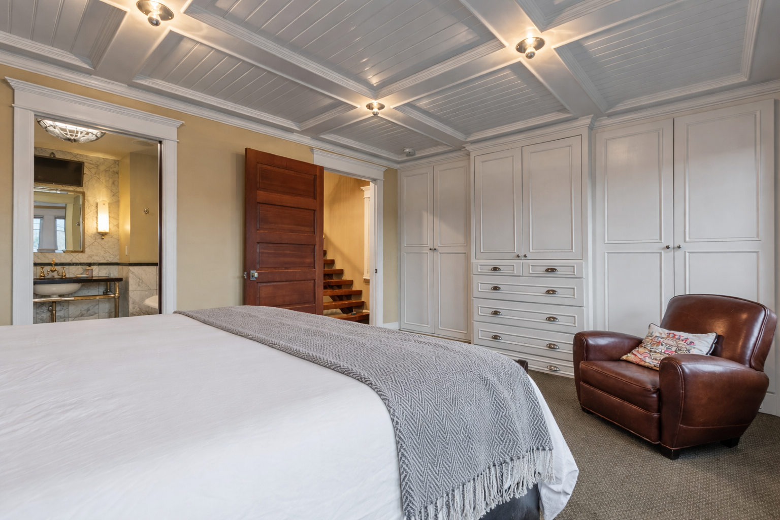 Unwind in your elegant bedroom featuring a cozy leather chair, beautiful coffered ceiling, and luxurious linens for ultimate comfort.