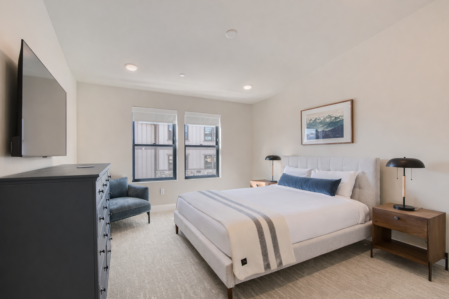 Stay in this serene, modern bedroom featuring a plush king-sized bed with a stylish channeled headboard. Large windows flood the space with light, while a cozy armchair offers the perfect spot to unwind.