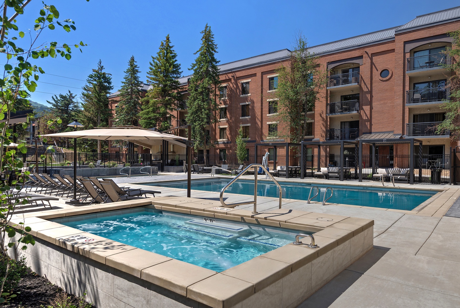 A sun-soaked courtyard awaits with sparkling pool and hot tub surrounded by tall pines, shaded loungers, and mountain views—perfect for lazy afternoons or evening soaks.