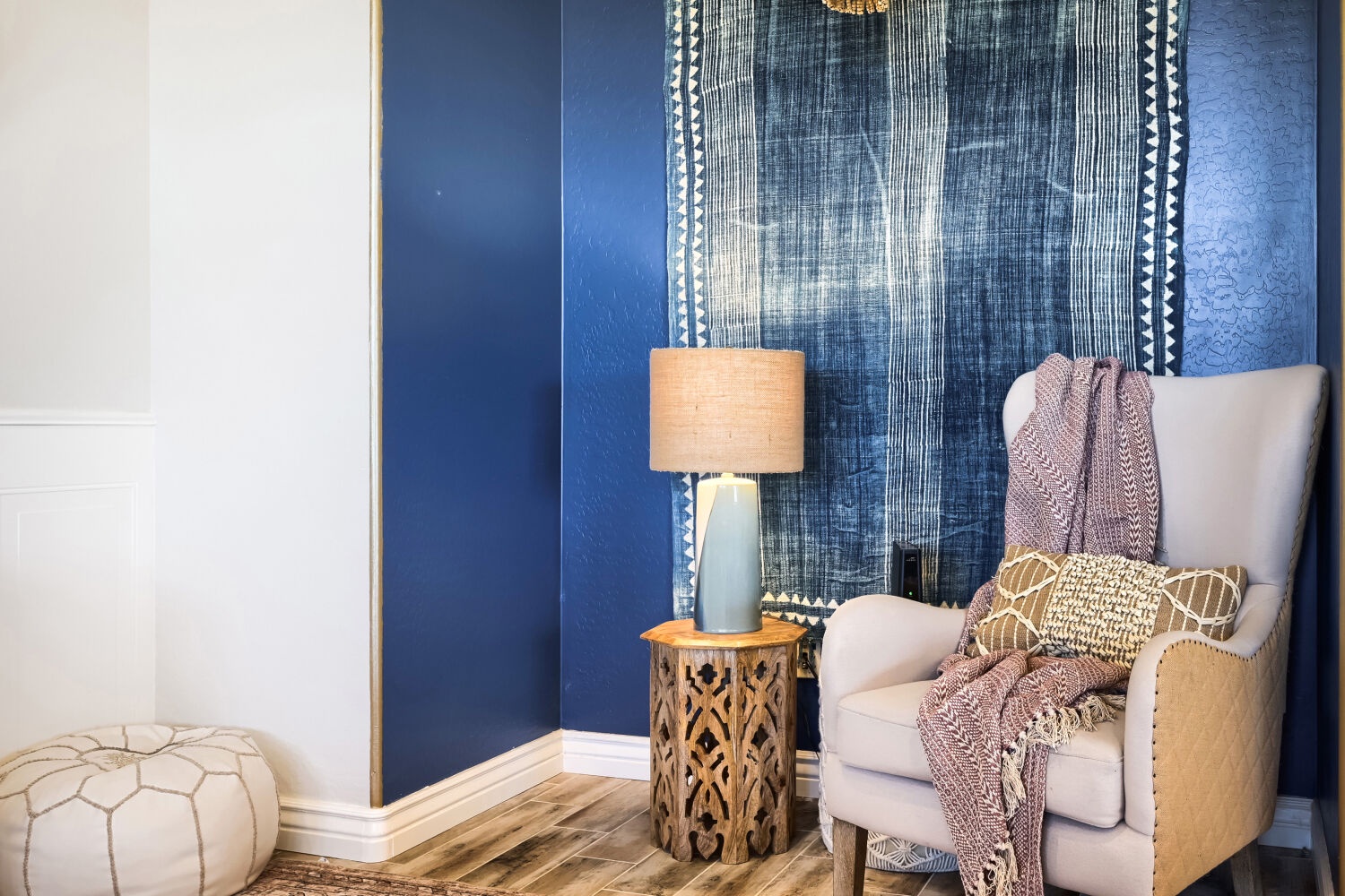 Your cozy reading corner awaits—settle into this plush armchair, wrap yourself in soft throws, and unwind beneath the striking indigo textile that sets a serene, artful mood.
