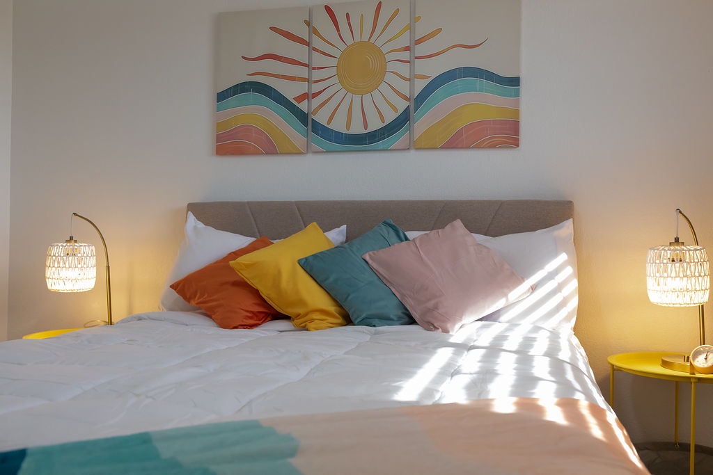 Wake up to cheerful sunshine streaming across your dreamy bed, where colorful pillows invite restful nights beneath vibrant wall art.