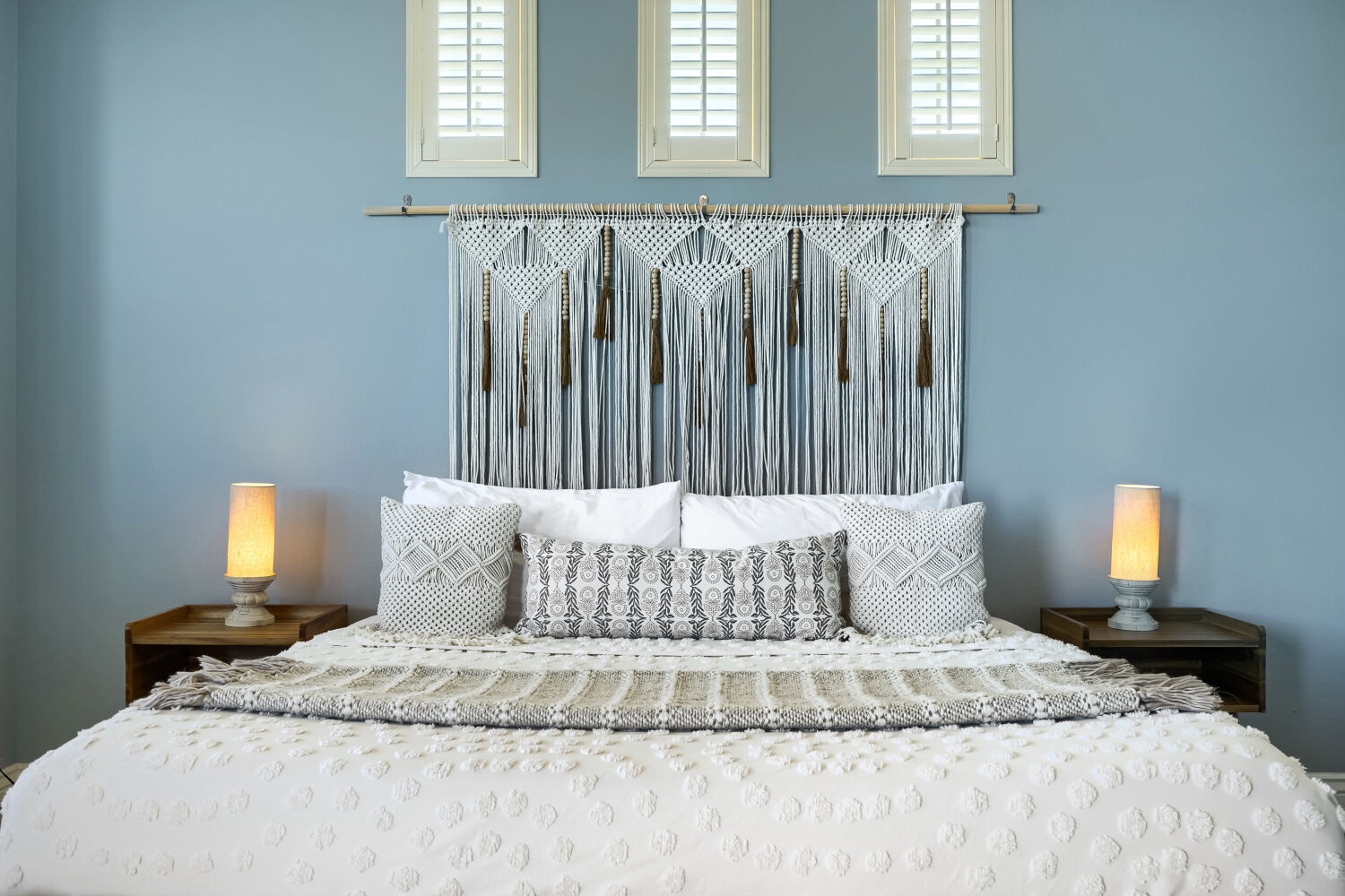Sink into this dreamy bed with handcrafted macramé headboard and soft layered linens. Natural light streams through shuttered windows, illuminating your serene coastal-style retreat where restful nights await.