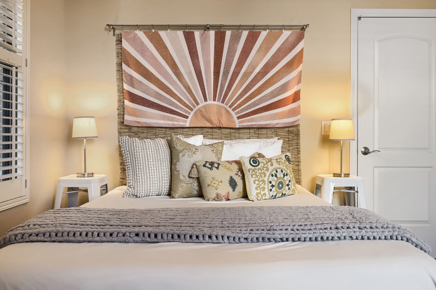 Your serene bedroom awaits with a plush bed, warm lighting, and a unique woven headboard framing a sunrise tapestry that brings calm and inspiration.