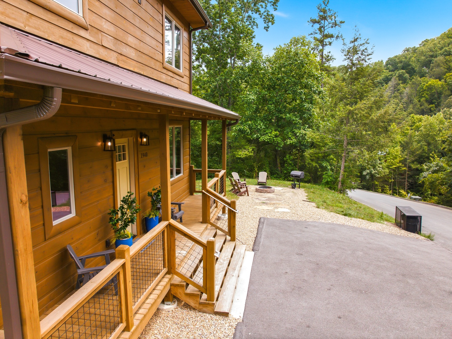 This charming wood-sided cabin is nestled among lush trees in the surrounding natural landscape, with a welcoming entrance and spacious parking area. The property features a fire pit and outdoor seating area perfect for gathering under the open sky.