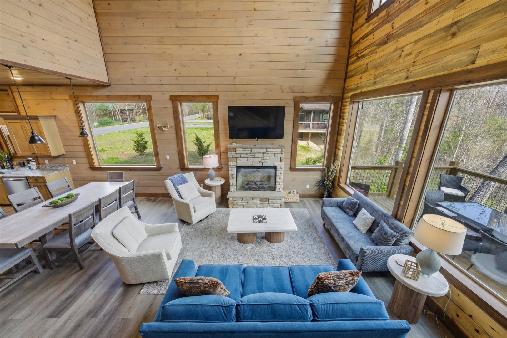 Relax in your spacious open-concept living area featuring warm wood walls, a cozy fireplace, and stunning forest views through panoramic windows.