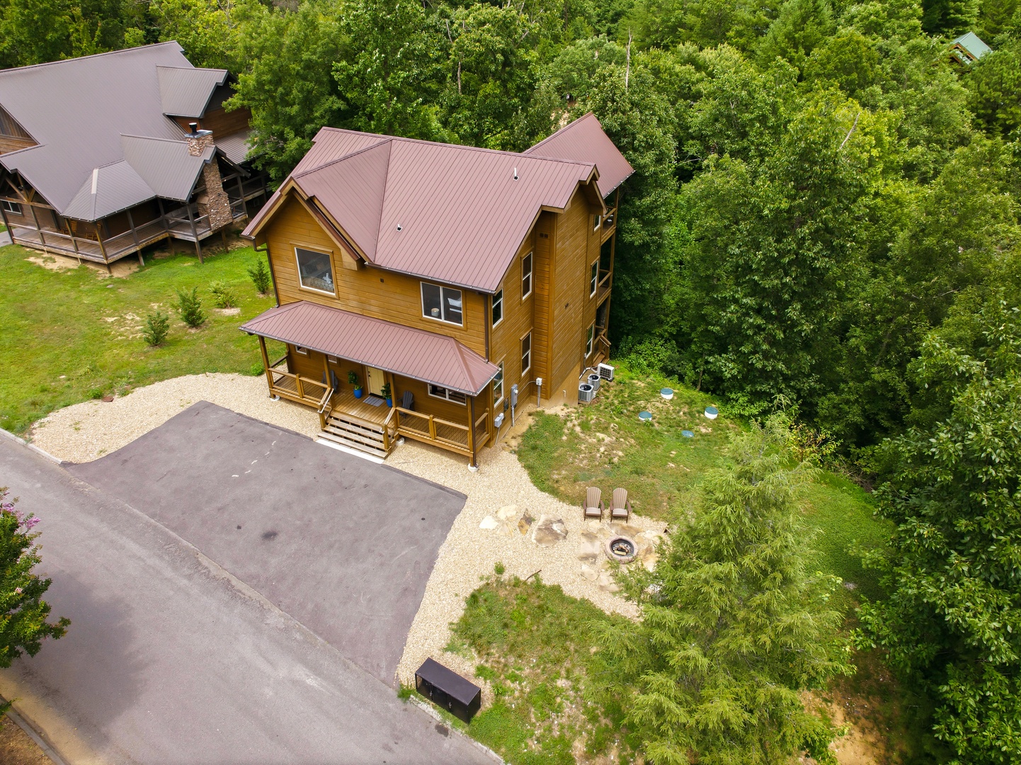 Aerial view of a charming two-story cabin nestled among lush forest surroundings, featuring dedicated parking, outdoor fire pit seating, and covered front porch.