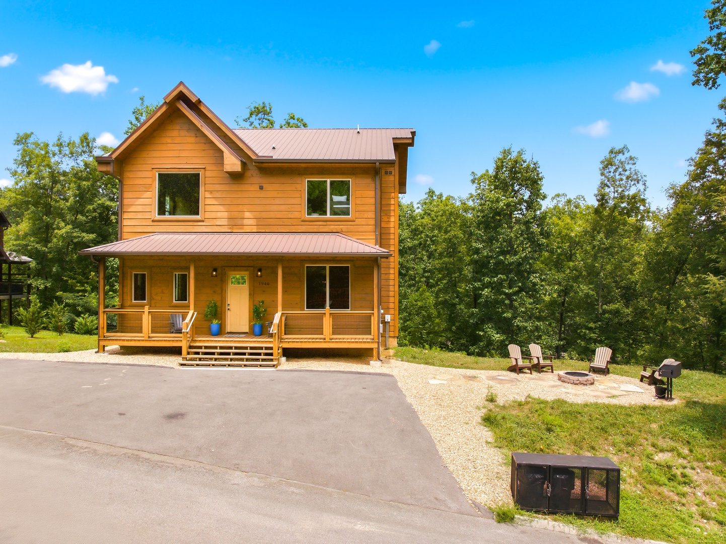 A warm two-story cabin with a covered front porch and fire pit area sits nestled among mature trees, offering easy parking and plenty of outdoor space for relaxing under the open sky.