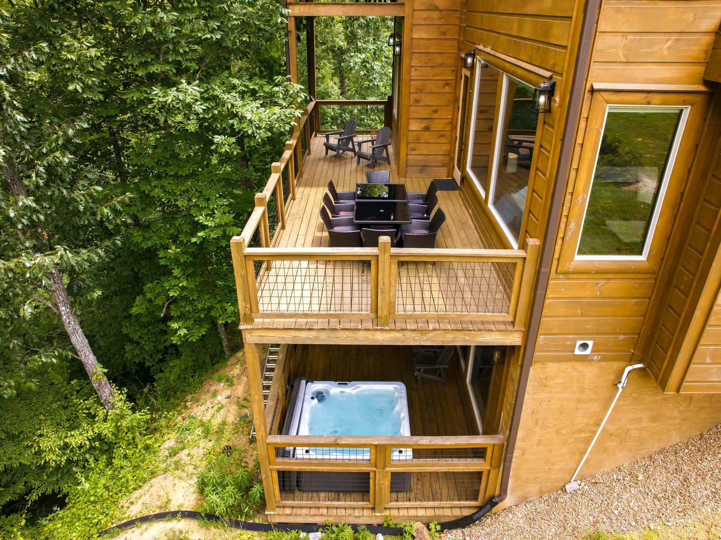 An aerial view of a private cabin retreat nestled among trees, featuring multi-level wooden decks, a secluded hot tub, and outdoor dining space surrounded by lush greenery.