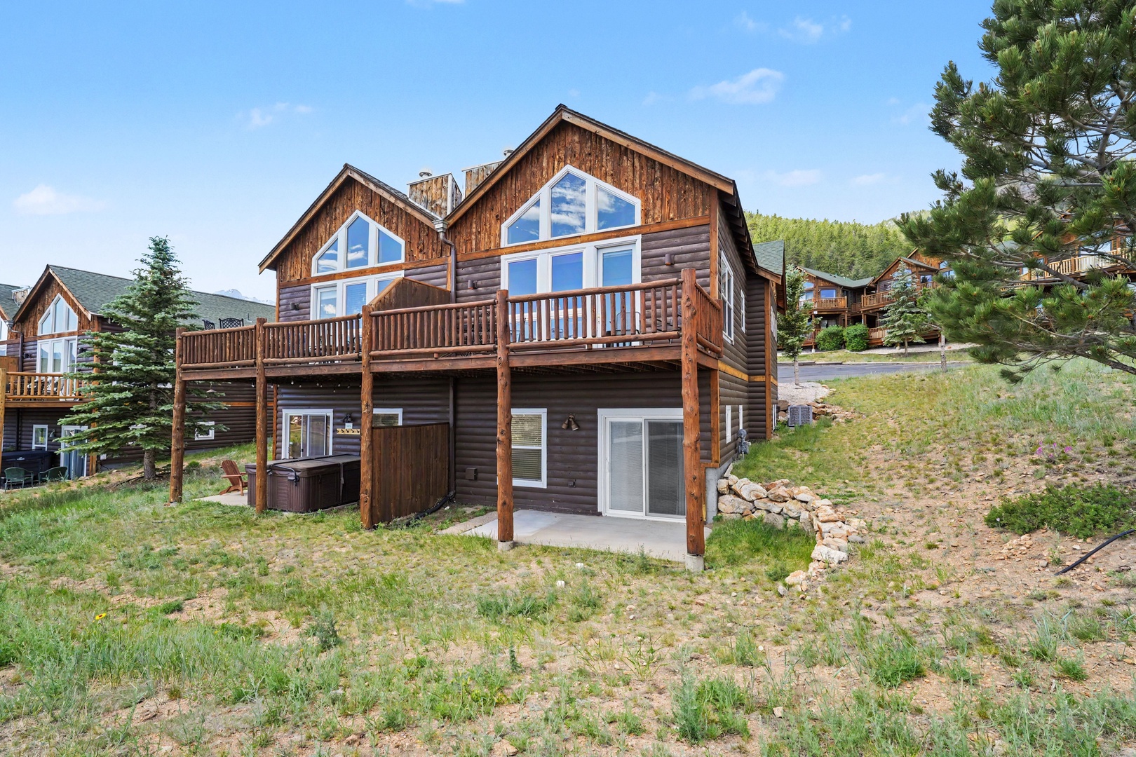 A charming mountain condo featuring wood siding, large windows, spacious decks, and a hot tub, nestled among evergreens in a peaceful neighborhood setting.