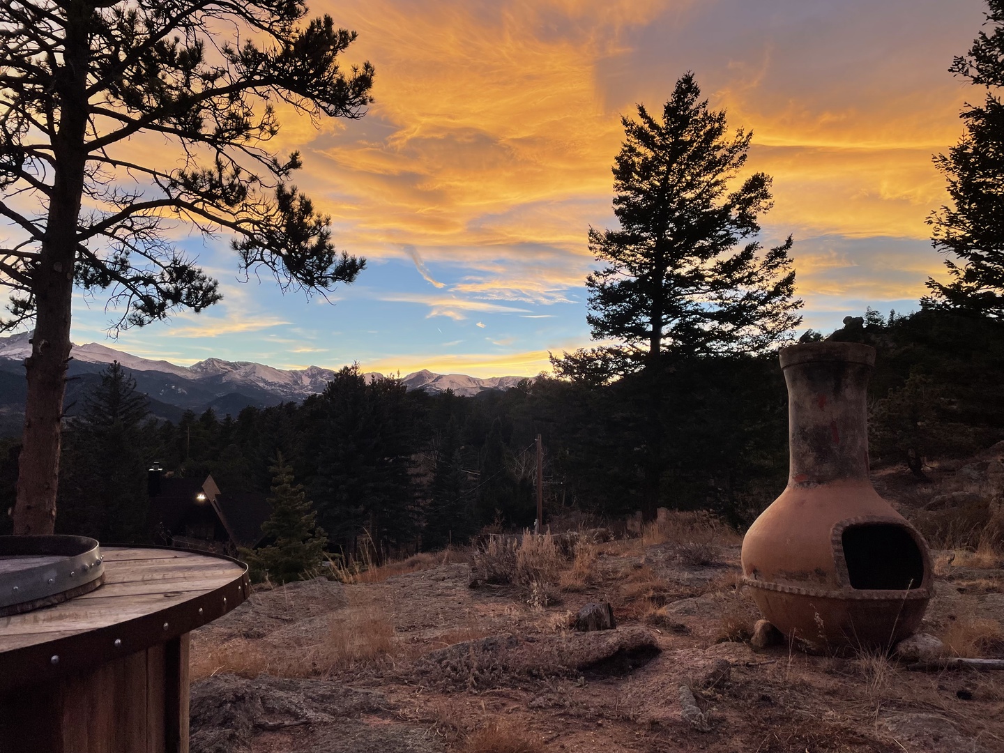 Golden sunset paints the sky over mountain peaks with a seating area framed by towering pines.