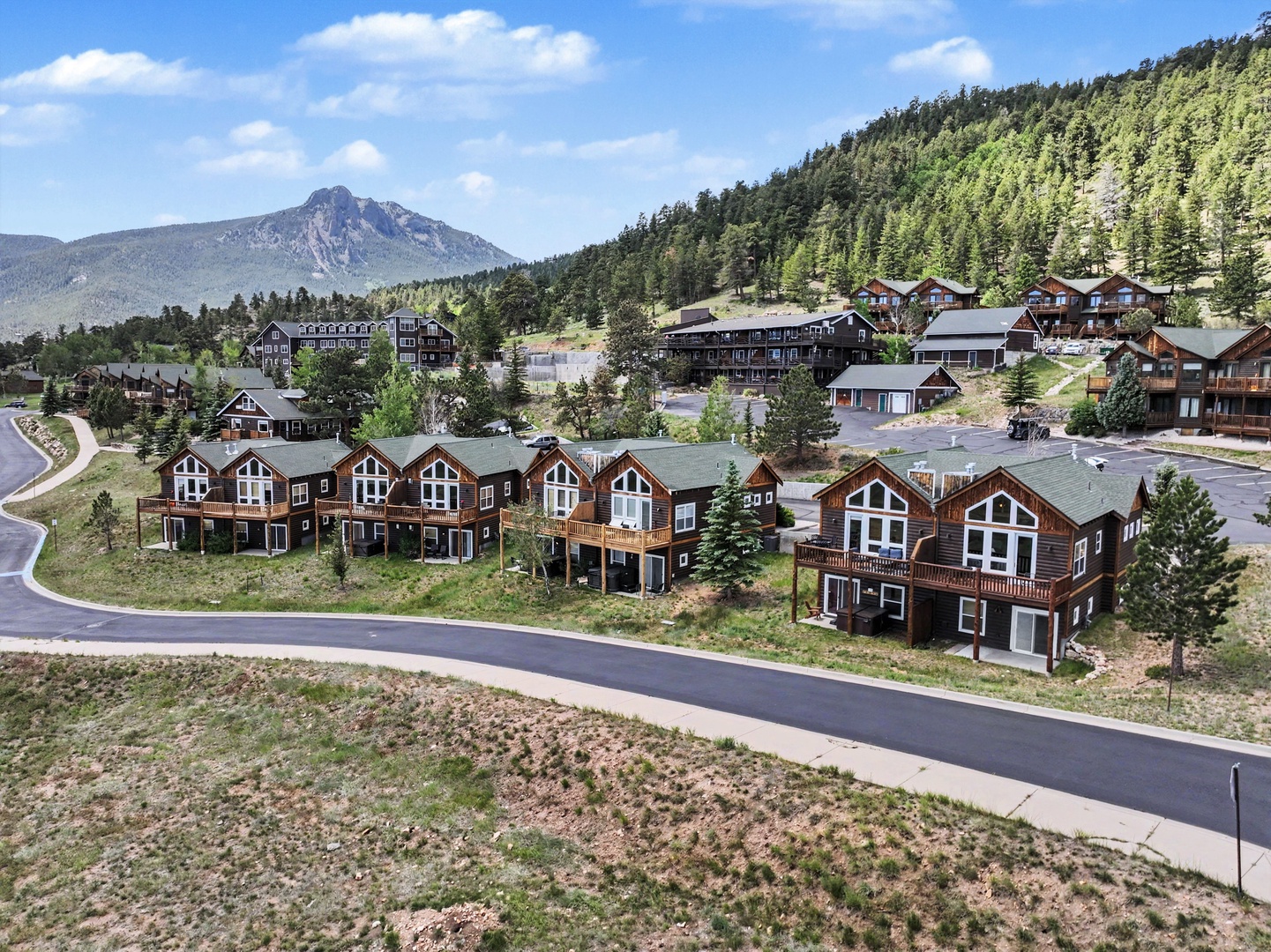 An elevated aerial view captures a scenic mountain resort community surrounded by forested hillsides and distant peaks, highlighting the property's picturesque location.