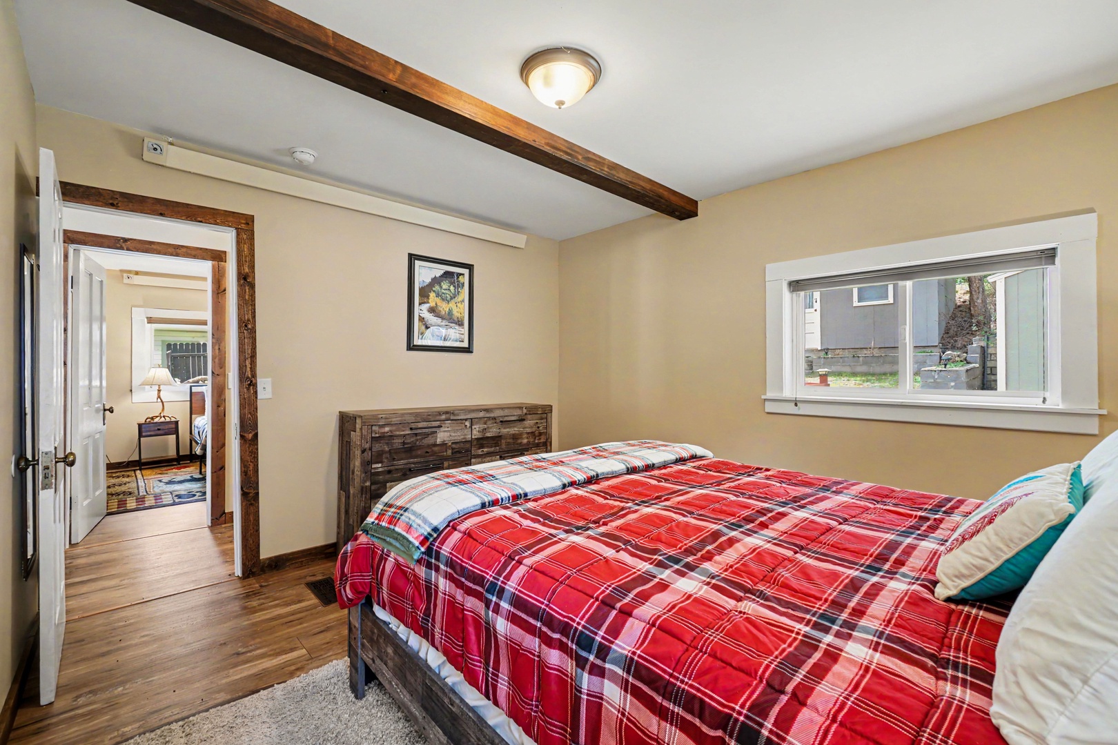 Your cozy bedroom features rustic wood-beam accents, a plush bed with cheerful plaid linens, and warm natural light—perfect for a peaceful retreat.