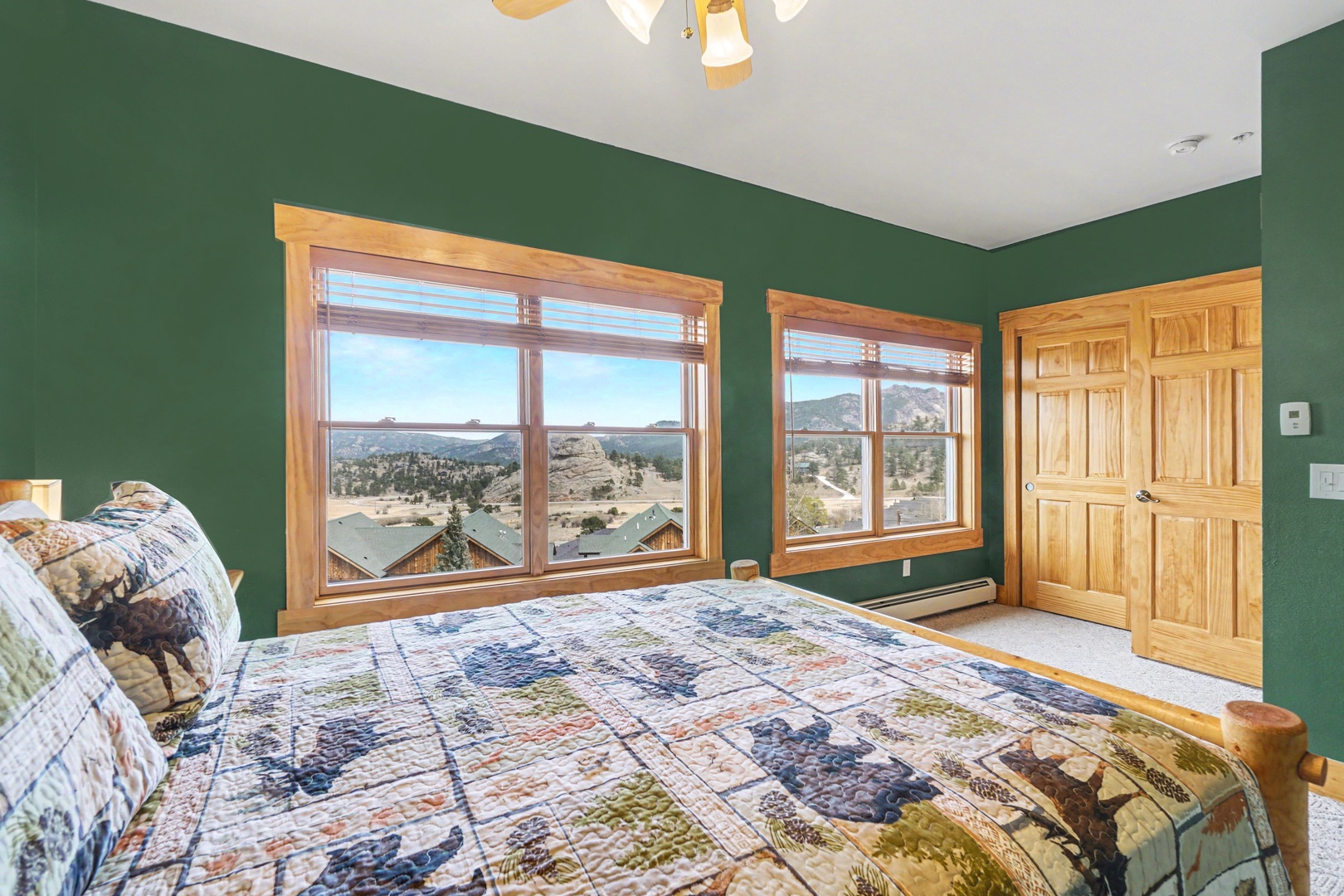 Wake up to breathtaking mountain views from your cozy bedroom's large windows, where rustic quilted bedding meets forest green walls.