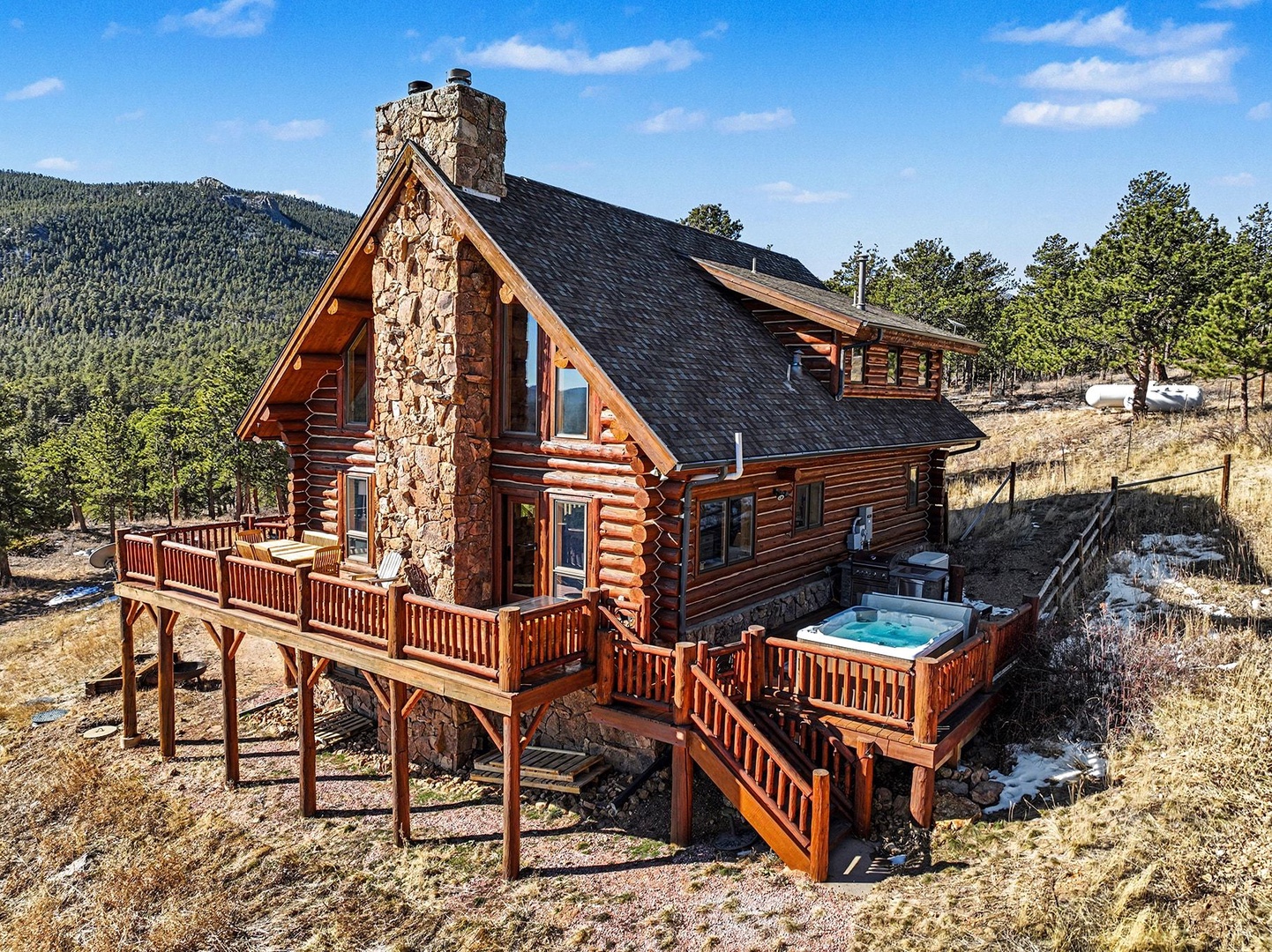Rustic log cabin with stone chimney nestled in mountain landscape, featuring wraparound deck and hot tub amid natural surroundings.