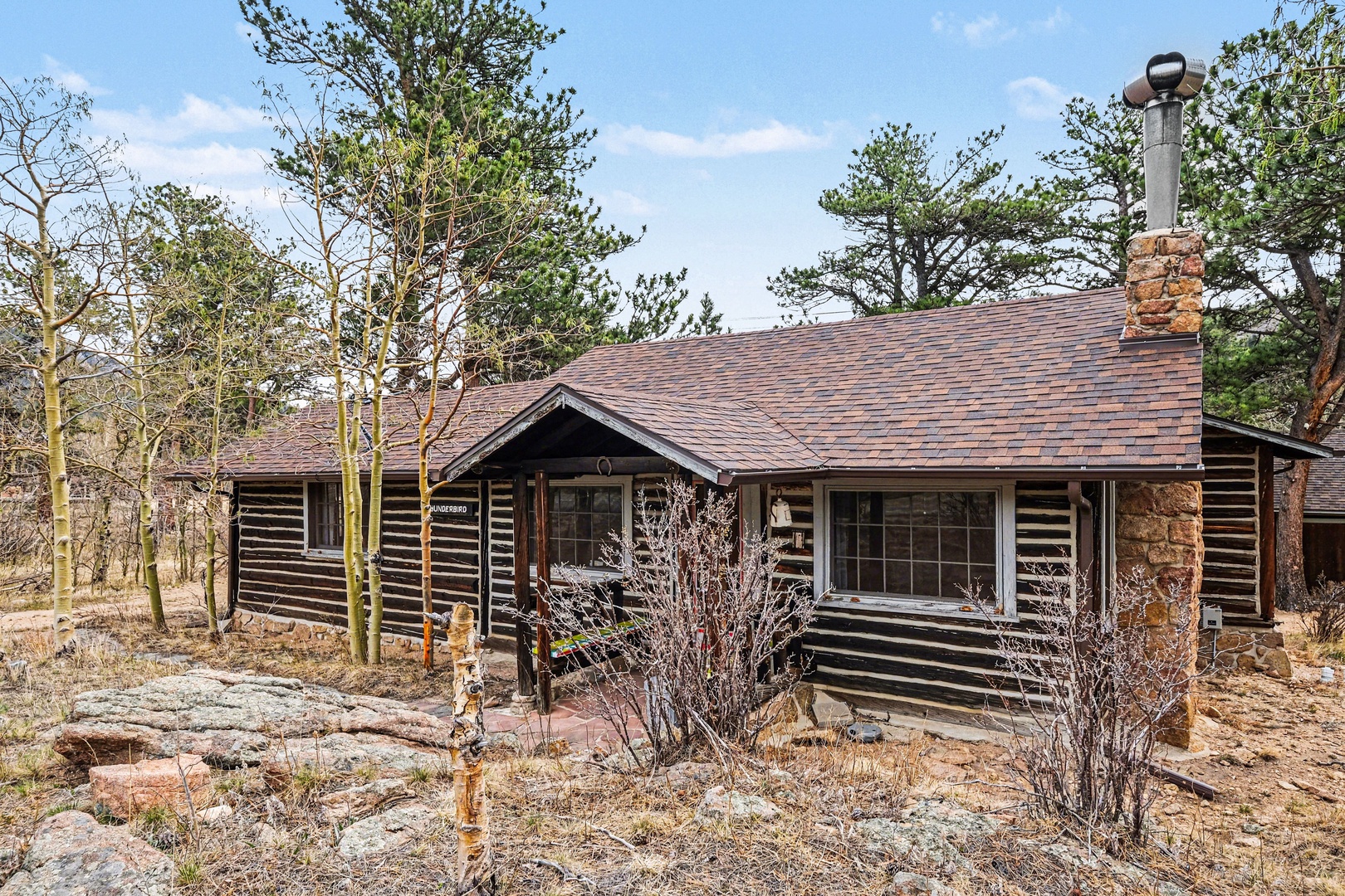 Rustic log cabin nestled among towering pines and natural rock formations, featuring classic stone chimney and peaceful woodland setting.