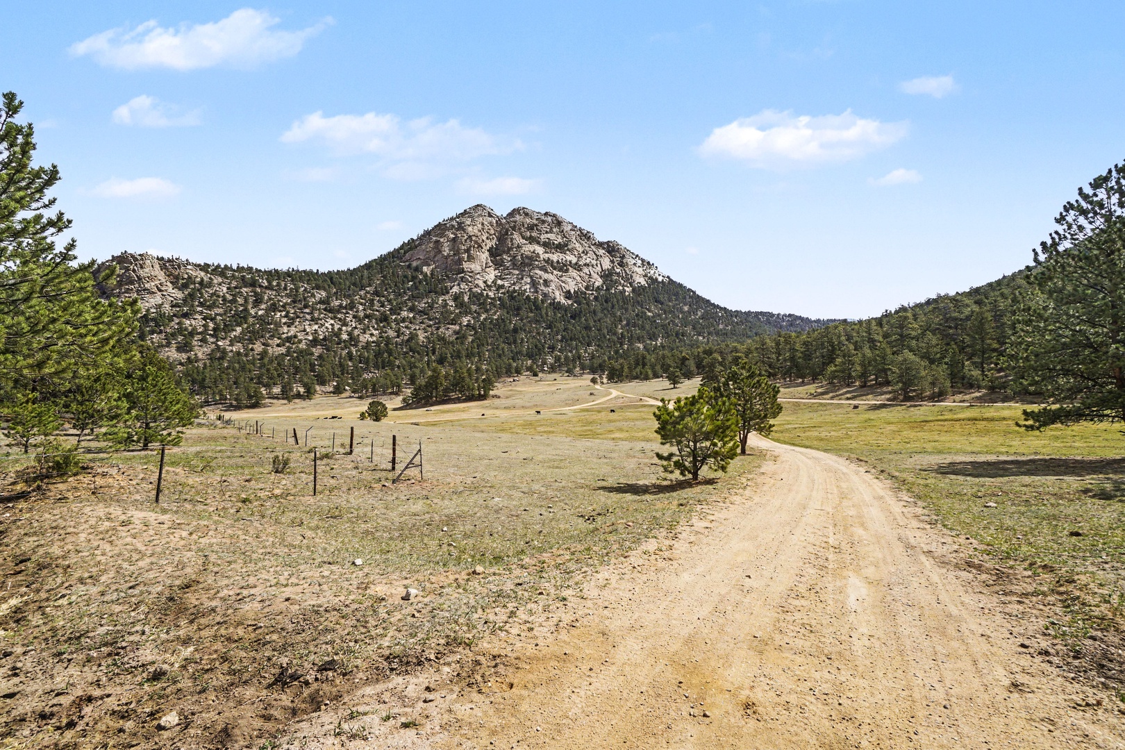 Scenic mountain landscape with winding dirt road through open countryside, surrounded by forested hills and expansive valley views.
